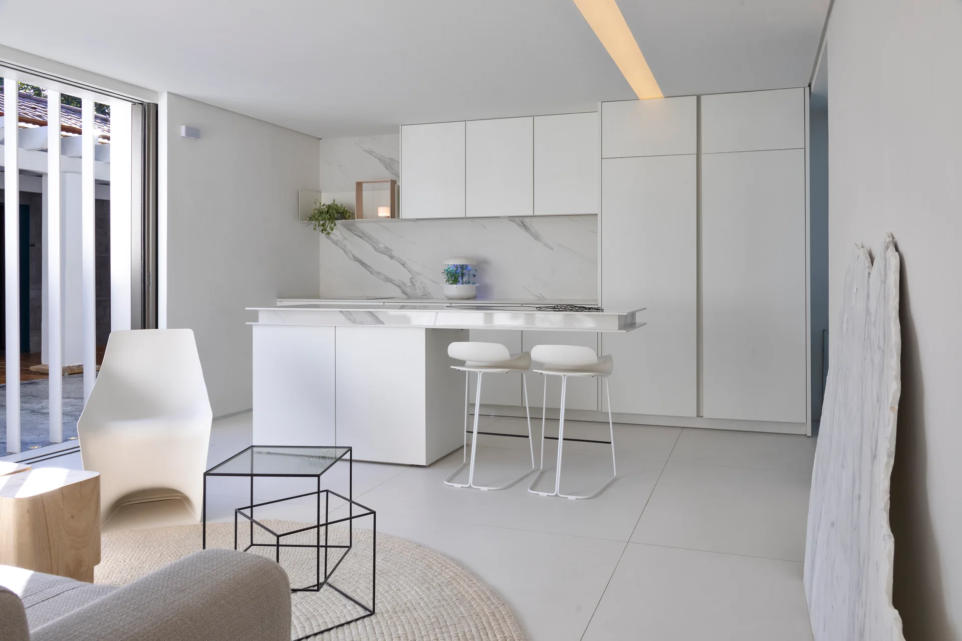 A modern white kitchen features flat-front cabinetry, a veined marble backsplash and island, white bar stools, and a minimalist living area.