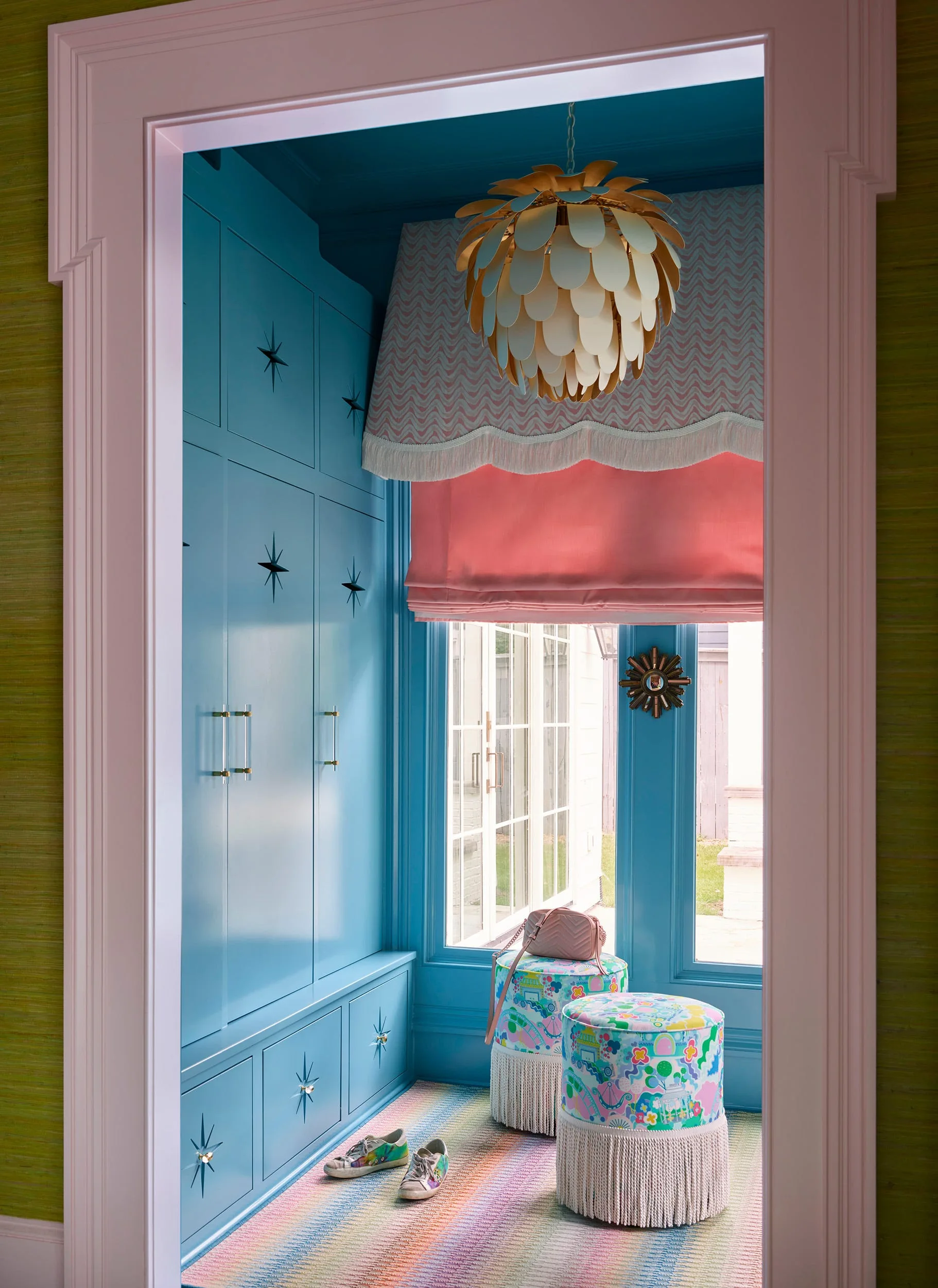 A small mudroom entryway painted entirely in sky blue features built-in cabinets with star-shaped hardware, fringed stools, and a pink window shade.