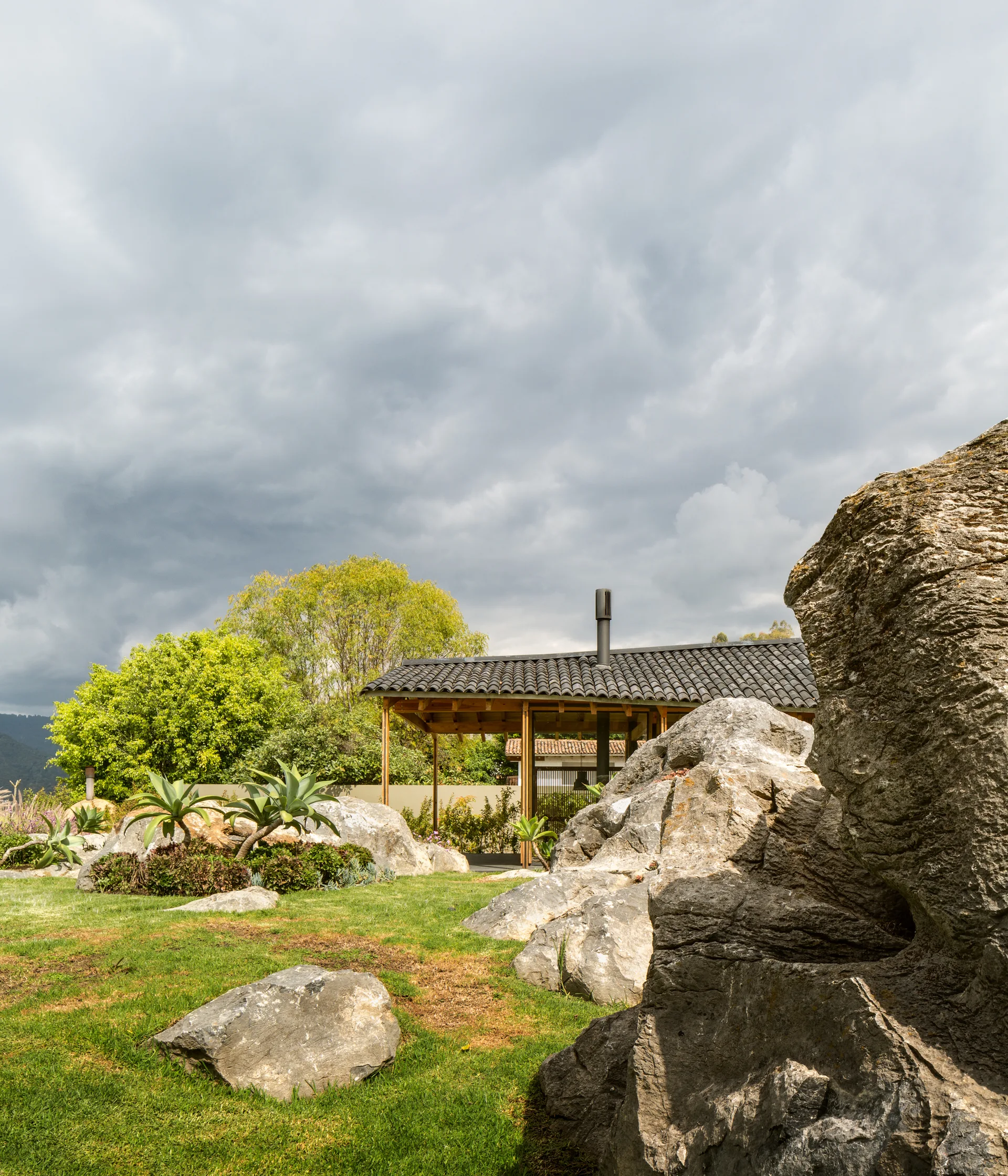 A green grassy lawn featuring large natural boulders and agave plants in front of an open-air pavilion with a dark tile roof.