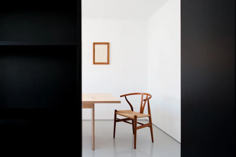 A wooden wishbone chair sits next to a light wood table in a minimalist white room framed by a dark foreground doorway.