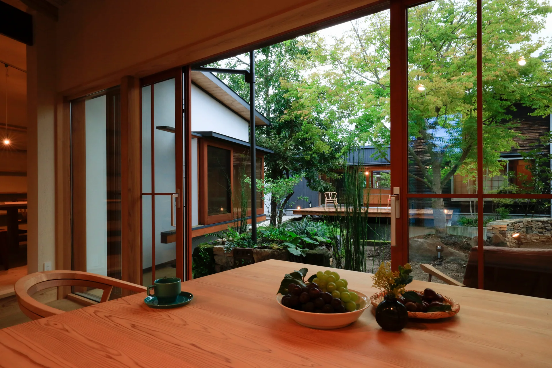 A smooth wooden dining table set with fruit bowls sits before a large glass window overlooking a green courtyard garden.