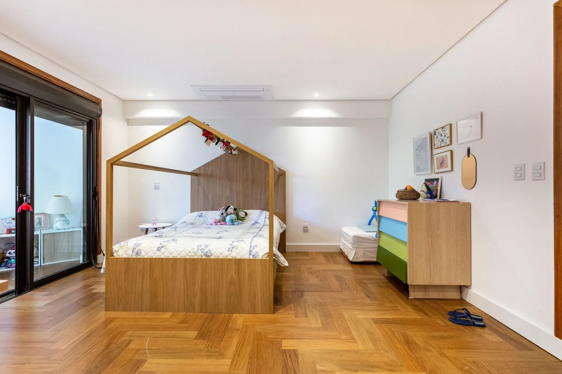 A children's bedroom featuring a house-shaped wooden bed frame, a colorful dresser, white walls, and herringbone parquet flooring.