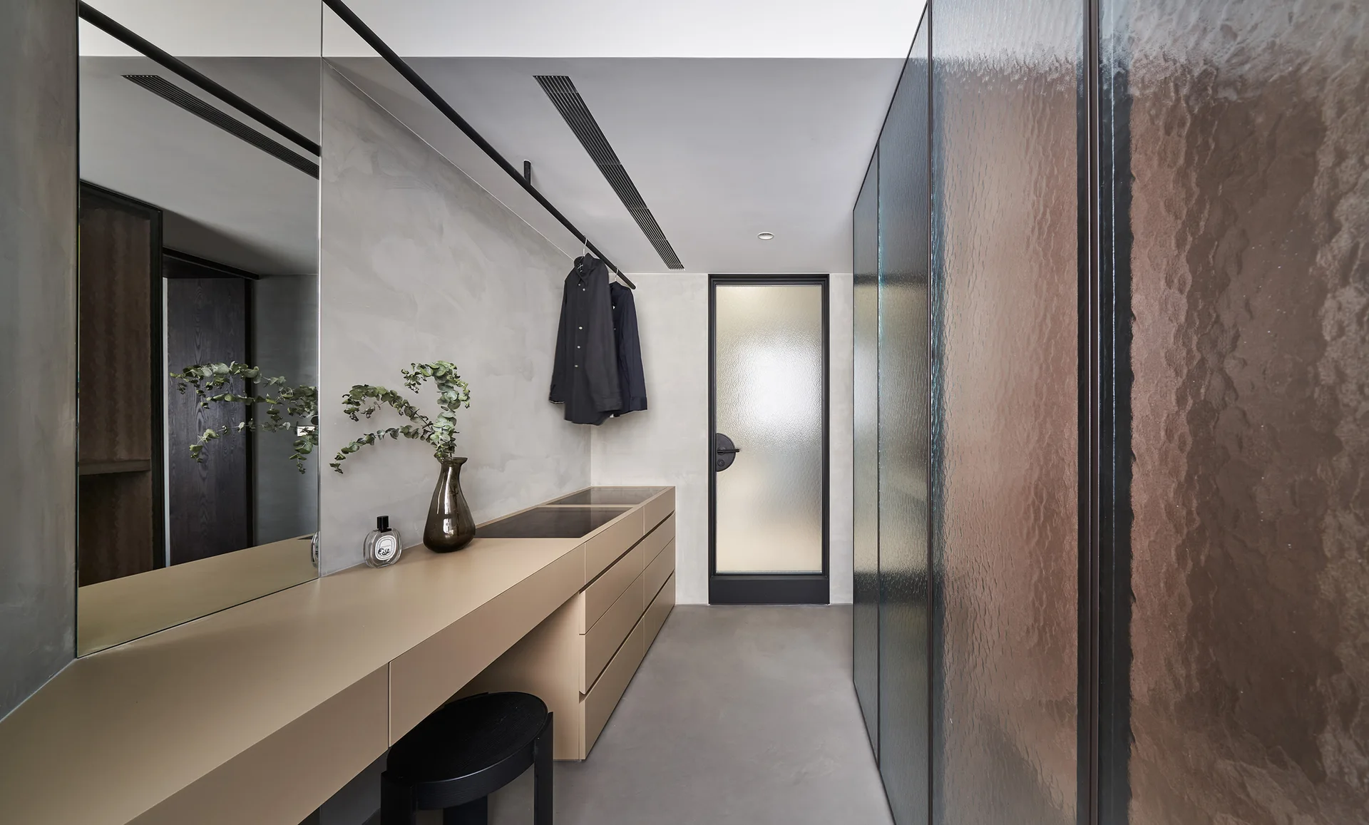A minimalist dressing room featuring a long beige vanity console with a black stool under a large mirror, opposite full-height textured glass closet doors.