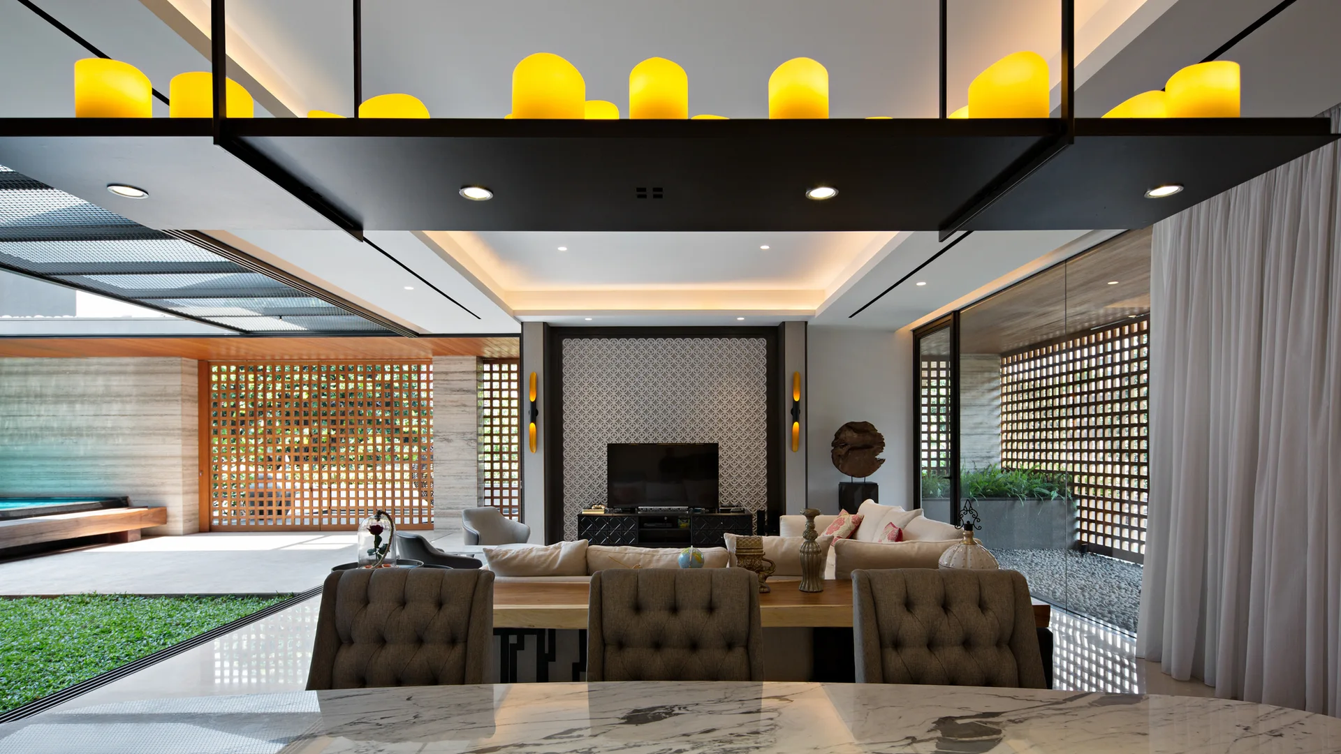 A modern open-plan living room features tufted grey dining chairs in the foreground, a beige sofa, and wooden lattice screens along the glass doors.