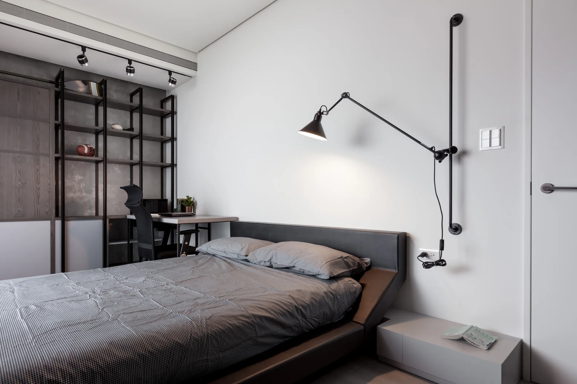 A modern bedroom featuring a low upholstered bed, a wall-mounted black swing-arm lamp, and a compact desk with dark metal shelving.