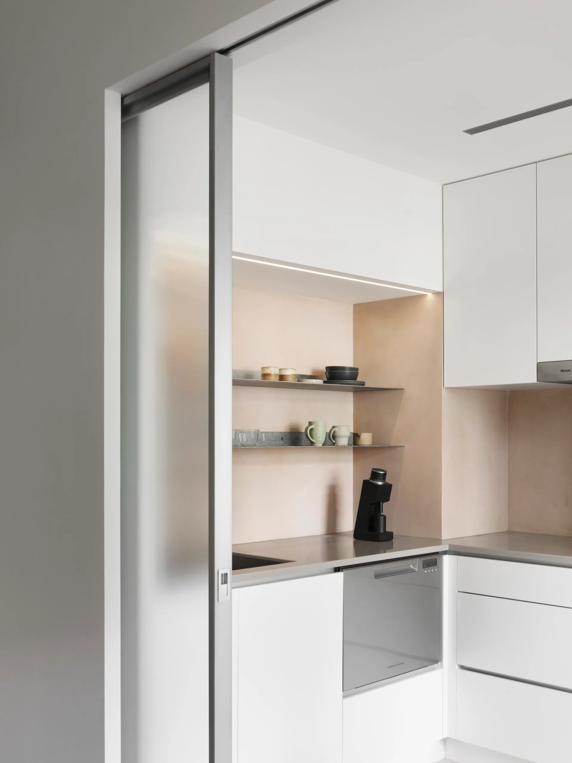 A minimalist white kitchen features a textured peach plaster backsplash with thin metal shelves behind a sliding frosted glass door.