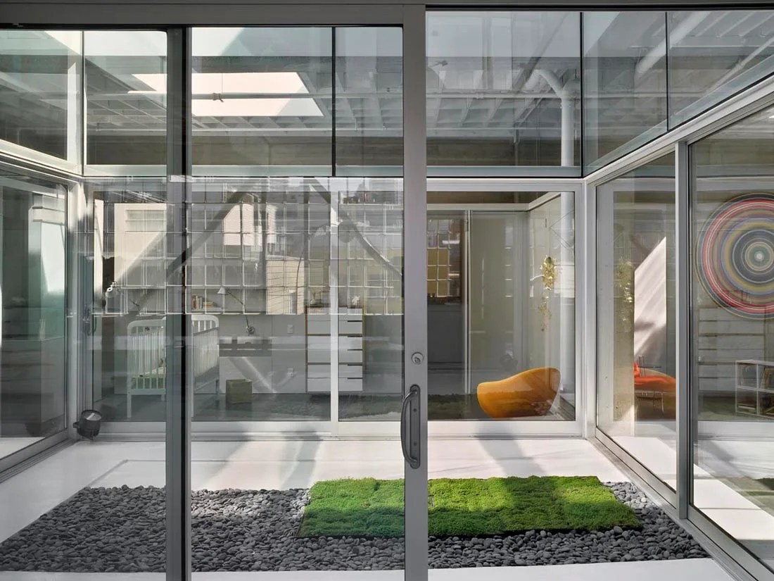 A modern glass-enclosed courtyard features dark grey pebbles and square green moss patches, with views into minimalist interior rooms.
