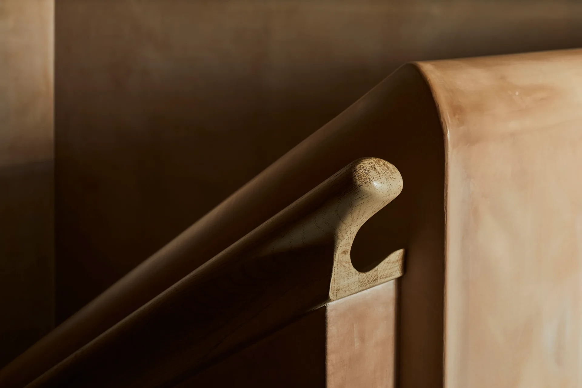 A close-up of a sculptural light wood handrail mounted against a curved matte plaster wall in warm earth tones with soft shadows.