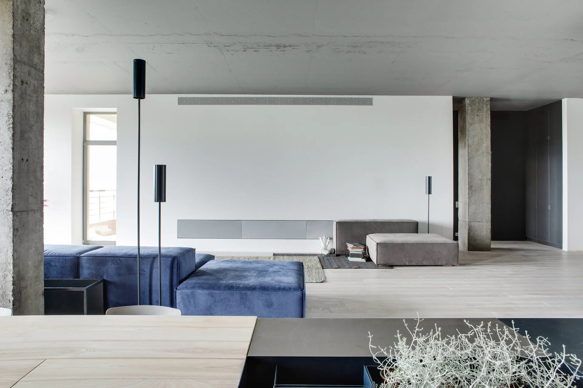 A minimalist living room features a blue velvet modular sofa, a raw concrete ceiling and column, and light wood flooring against a plain white wall.