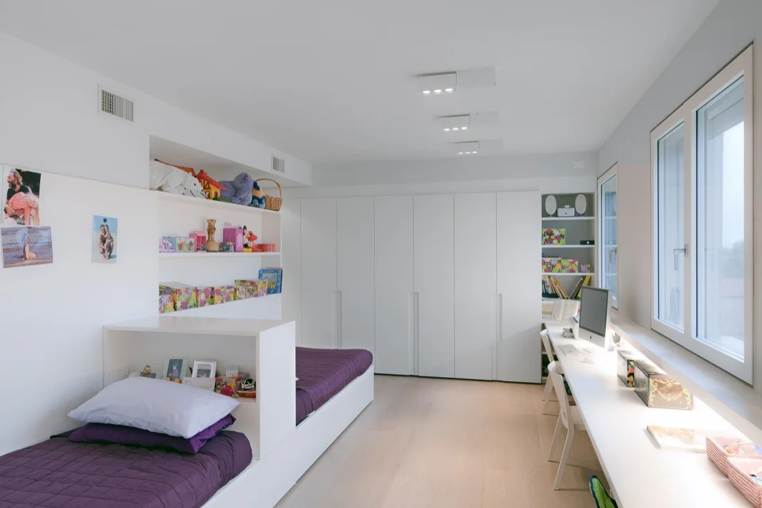A minimalist children's bedroom featuring twin beds with purple bedding, a long white desk beneath a window, and integrated white wardrobes.