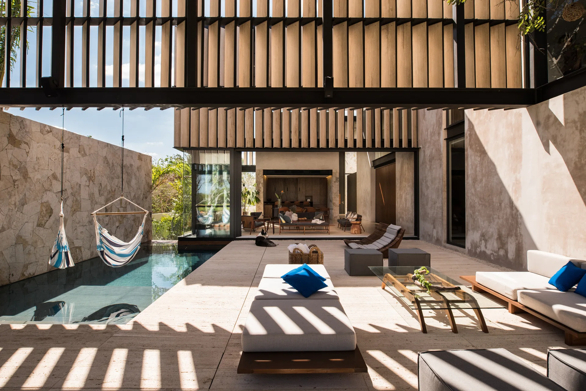 A modern outdoor patio features travertine stone tile flooring, low-profile gray seating, a hanging striped hammock chair, and a slatted wood ceiling.