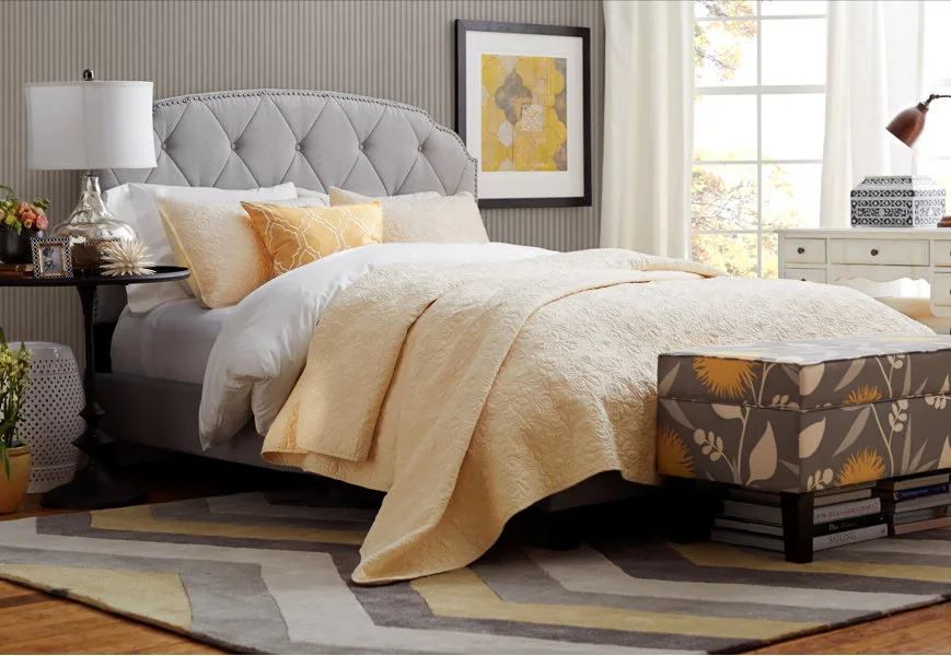 A grey tufted headboard bed is layered with yellow quilts next to a chevron patterned rug and floral print storage bench.
