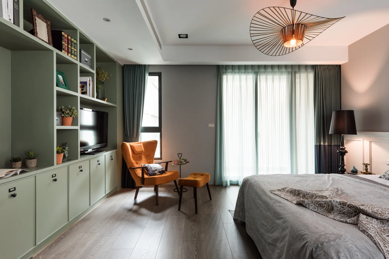 A modern bedroom features a sage green built-in shelving unit with a television, a burnt orange armchair with matching ottoman, and a bed.