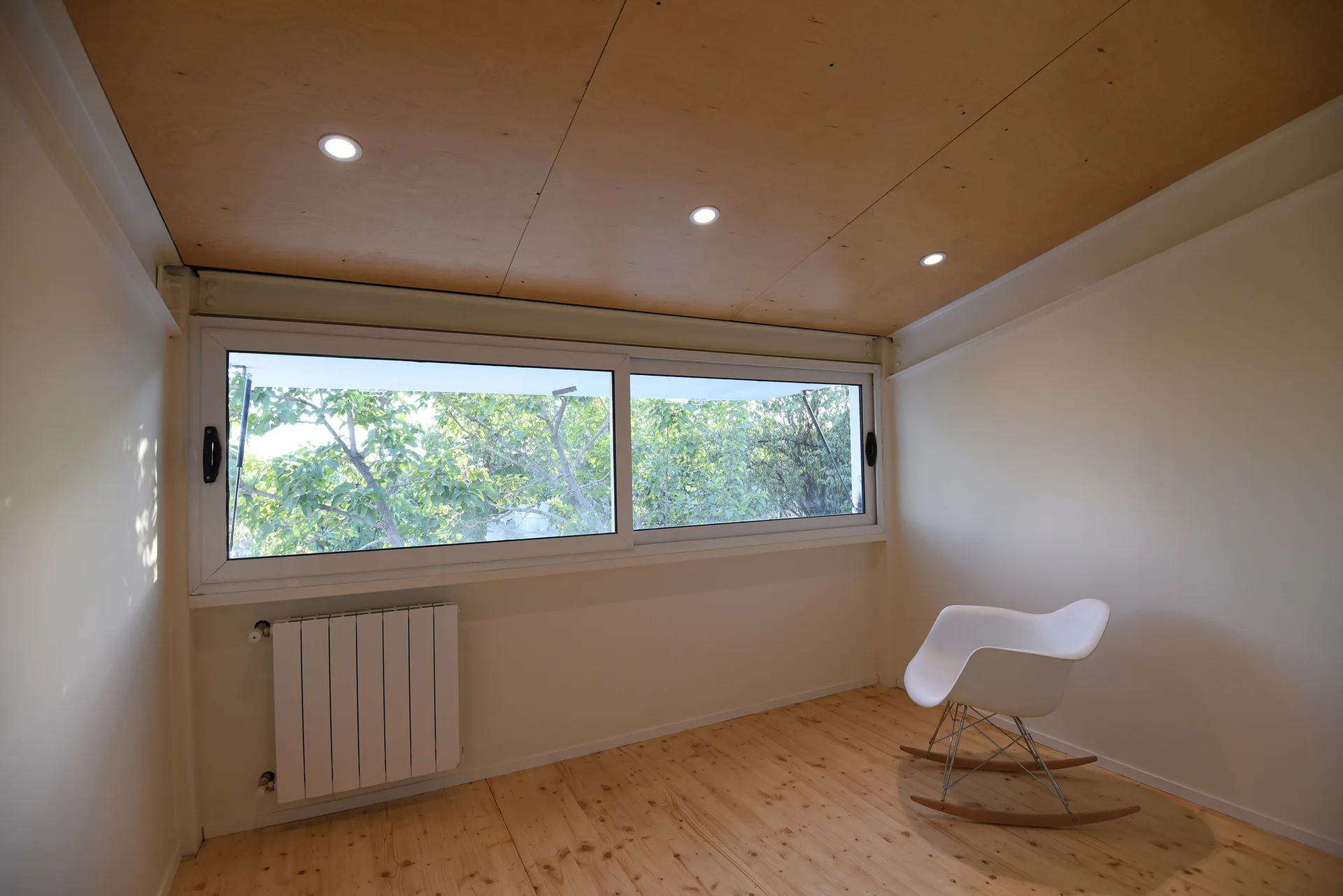 A minimalist room with light wood floors and ceiling, featuring a single white plastic rocking chair near a wide window displaying green trees.