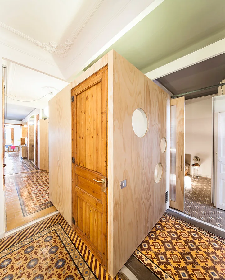 A modern plywood box partition sits in an apartment hallway featuring ornate patterned tile floors and historical white ceiling molding.
