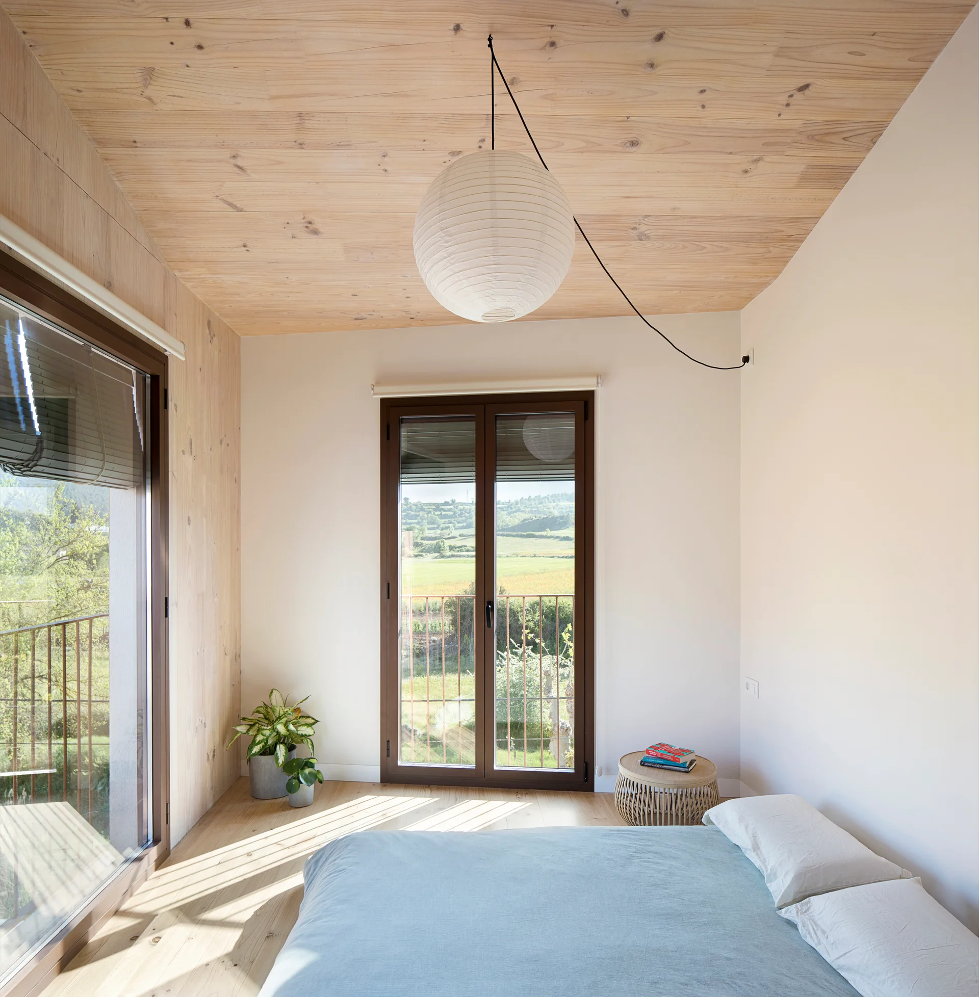 A minimalist bedroom features a low bed with blue linens, a wood-paneled ceiling, a white paper pendant lamp, and glass doors opening to a green valley.