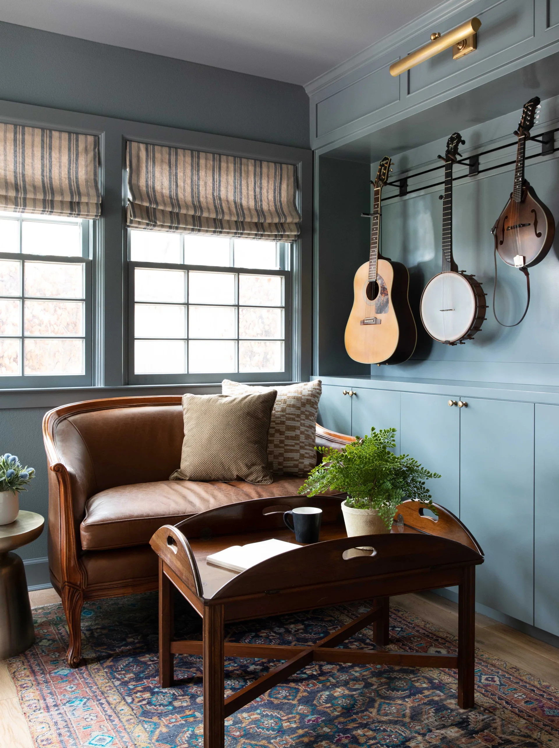 A brown leather settee sits below windows with striped roman shades next to slate blue cabinetry displaying wall-mounted musical instruments.