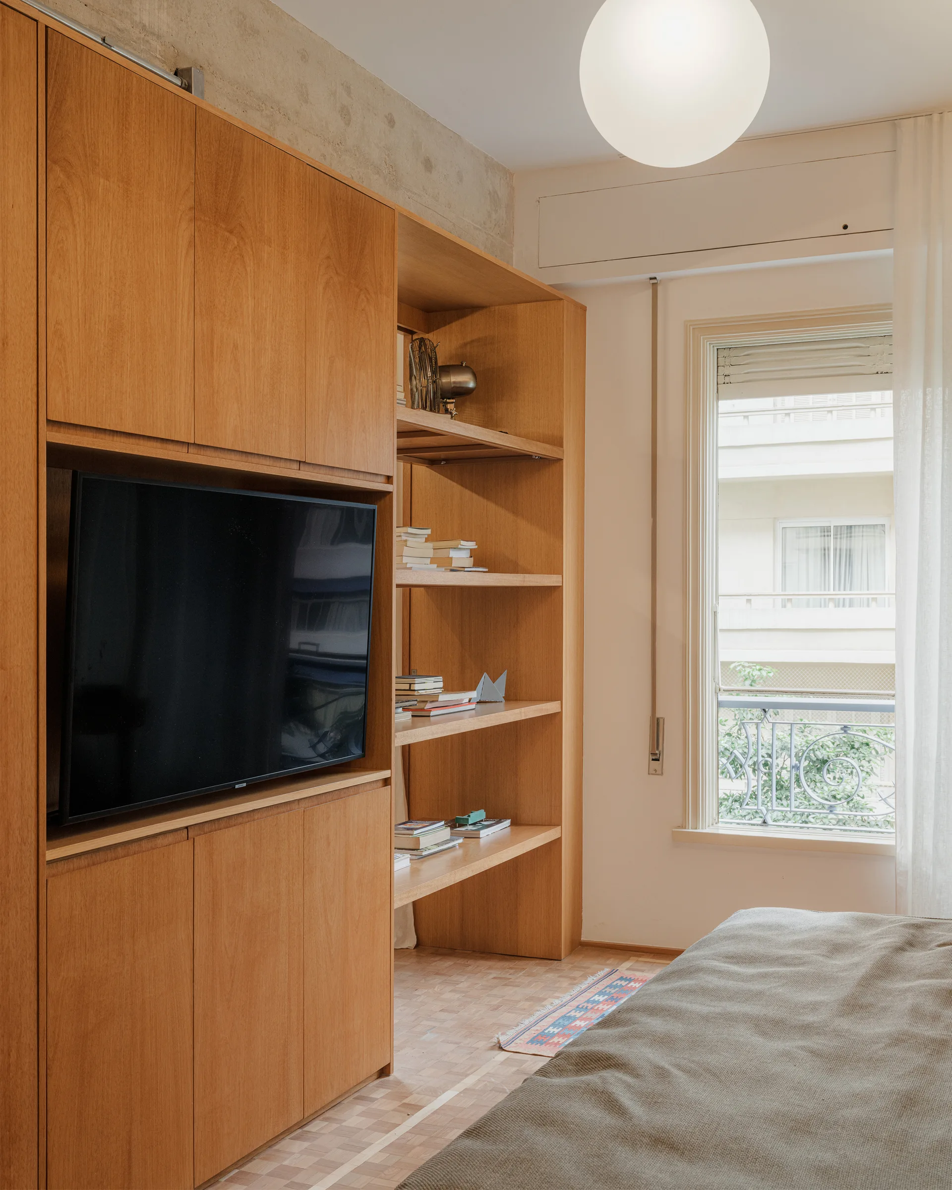 A bedroom features a custom light wood built-in unit containing a television and open shelving next to a window with sheer curtains.
