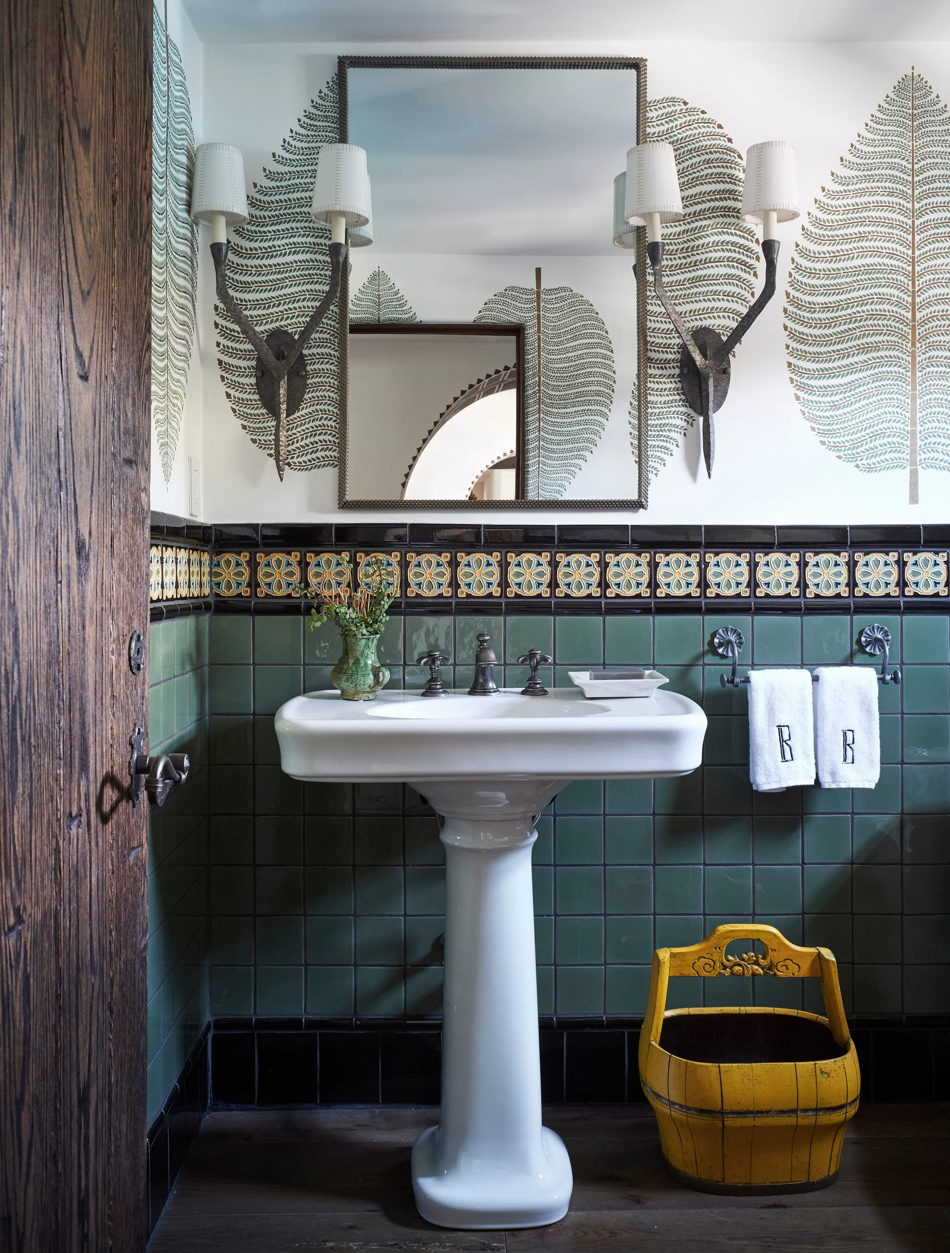 A bathroom features a white pedestal sink against glossy green wall tiles, botanical fern wallpaper, a rectangular mirror, and sconces.