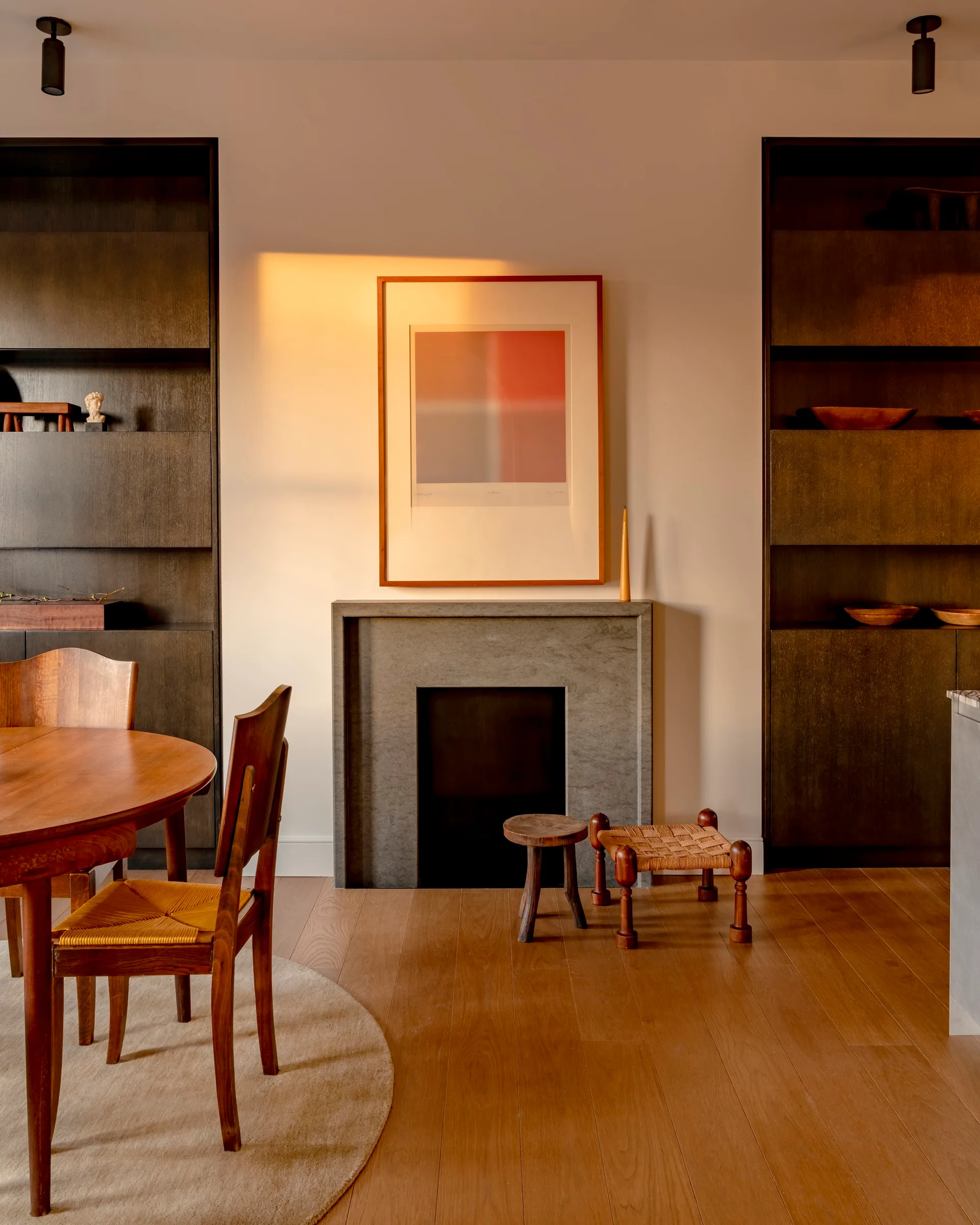 A stone fireplace with abstract framed art is flanked by tall dark wood built-in shelves in a sunlit dining area with wood furniture.
