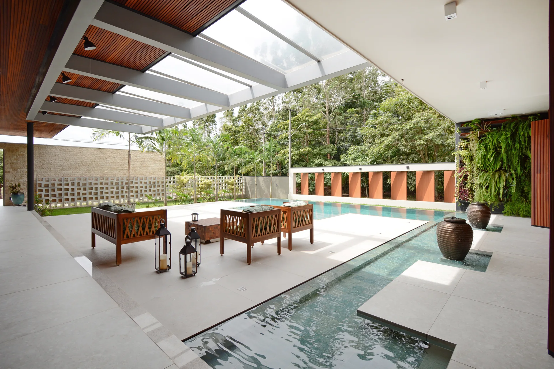 A covered outdoor patio features wooden armchairs, a swimming pool integrated into the tile floor, a large skylight, and a vertical green garden wall.