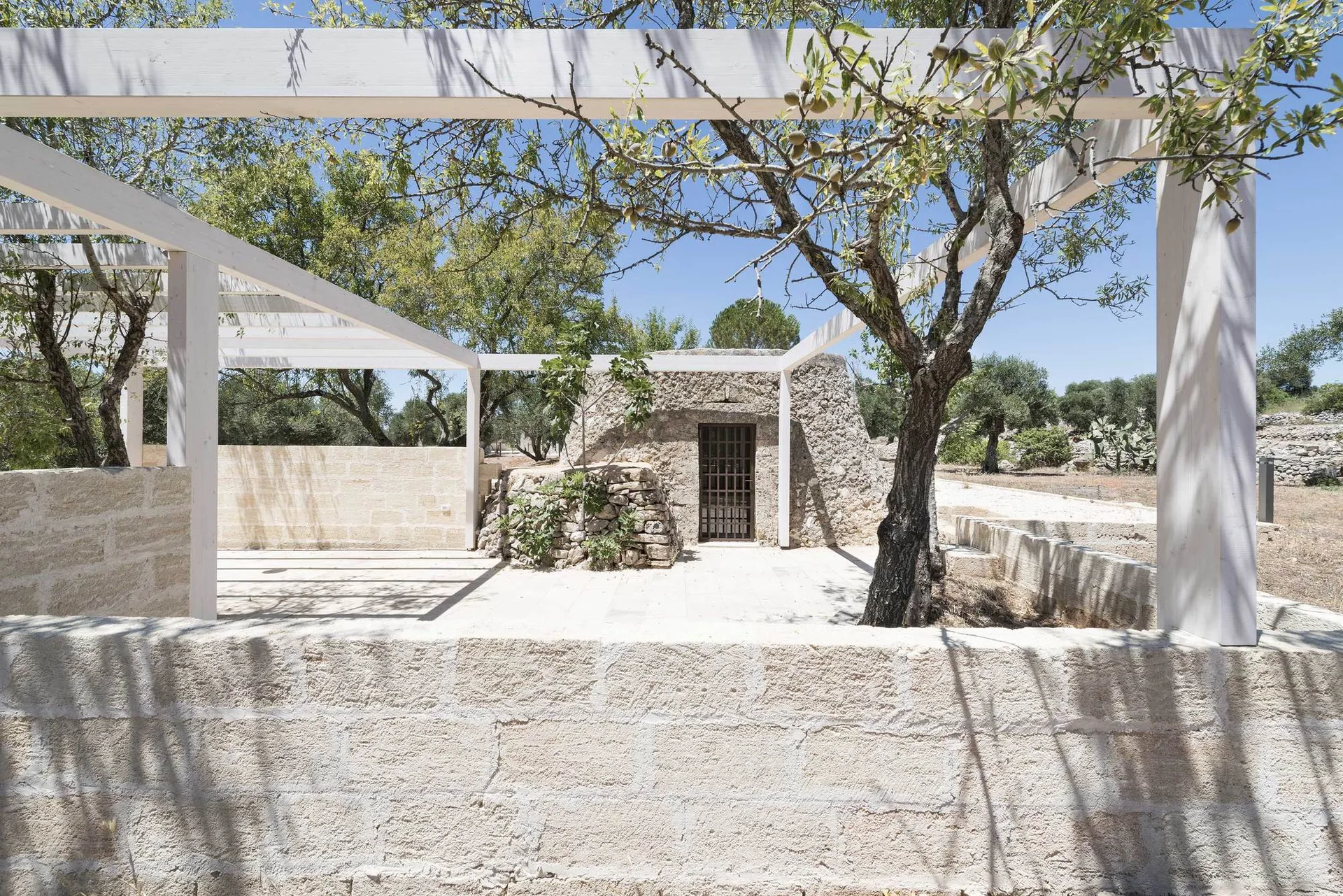 An outdoor stone patio features a minimalist white wood pergola framing a rustic circular stone hut and low limestone walls under a clear blue sky.