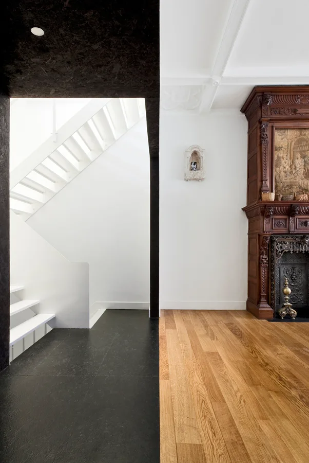A split room view shows a modern white staircase with black flooring next to a traditional room featuring light oak floors and an ornate wood fireplace.