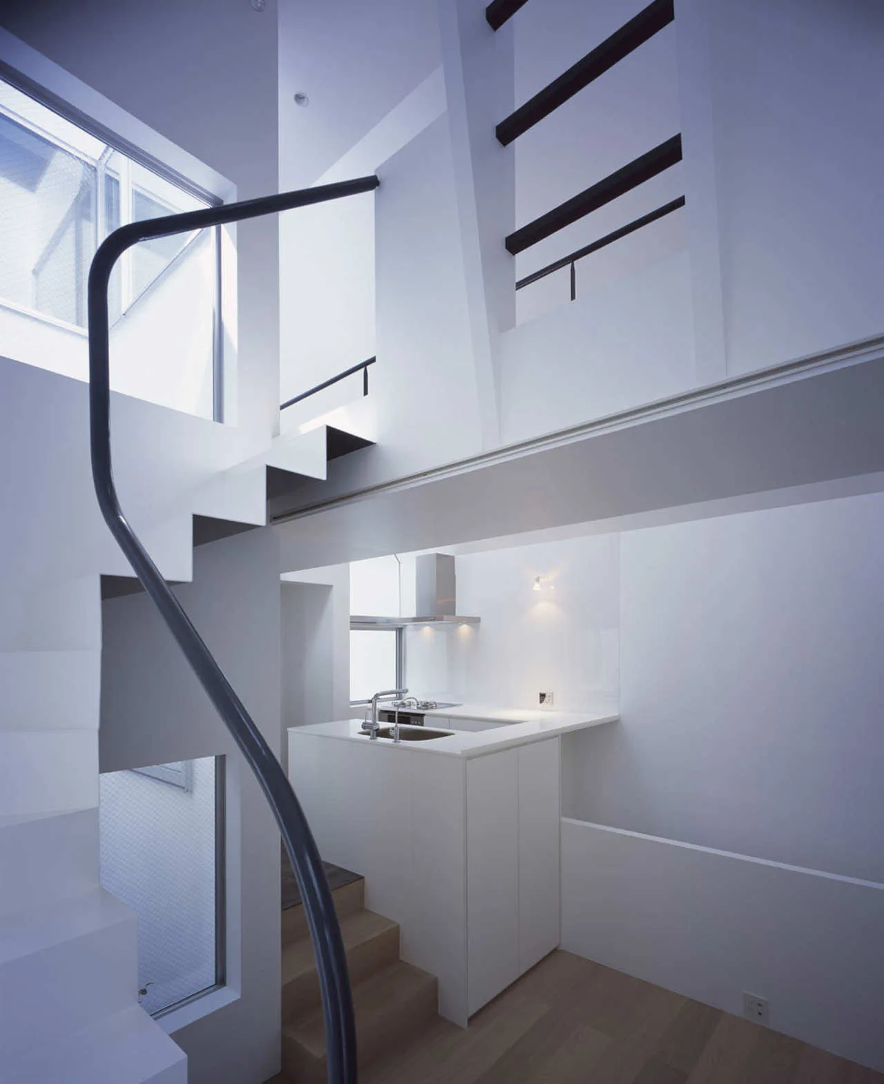 A minimalist white loft interior featuring a sculptural staircase with a black handrail and a compact kitchen beneath the upper level.