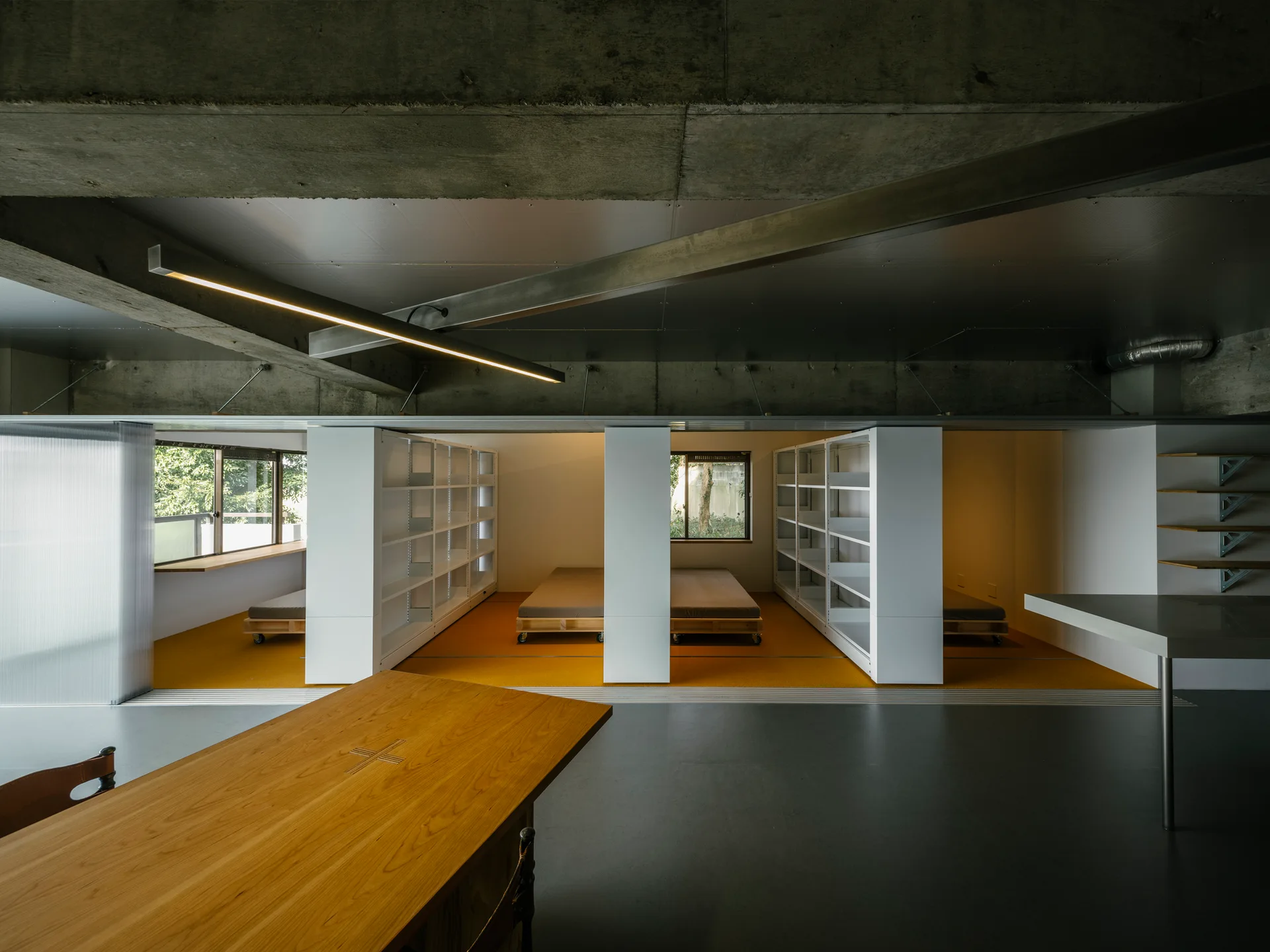An industrial room features an exposed concrete ceiling, a large wooden table, and modular white shelving units dividing sleeping areas with yellow flooring.