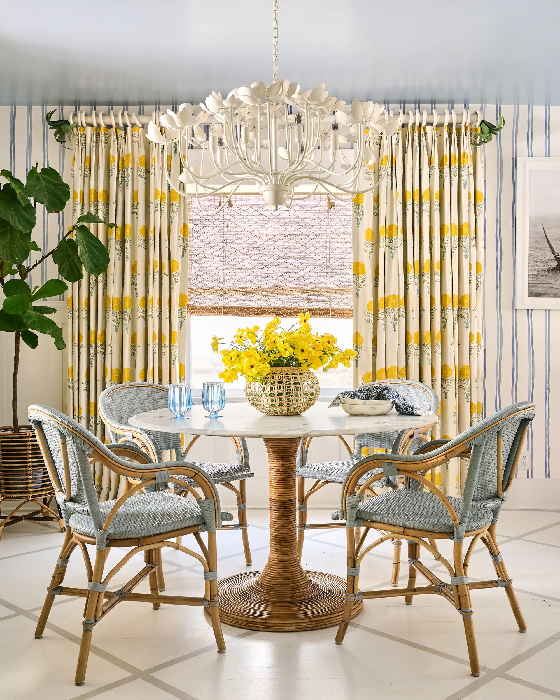 A round white marble dining table with a woven rattan base sits surrounded by blue wicker bistro chairs in a colorful striped room.