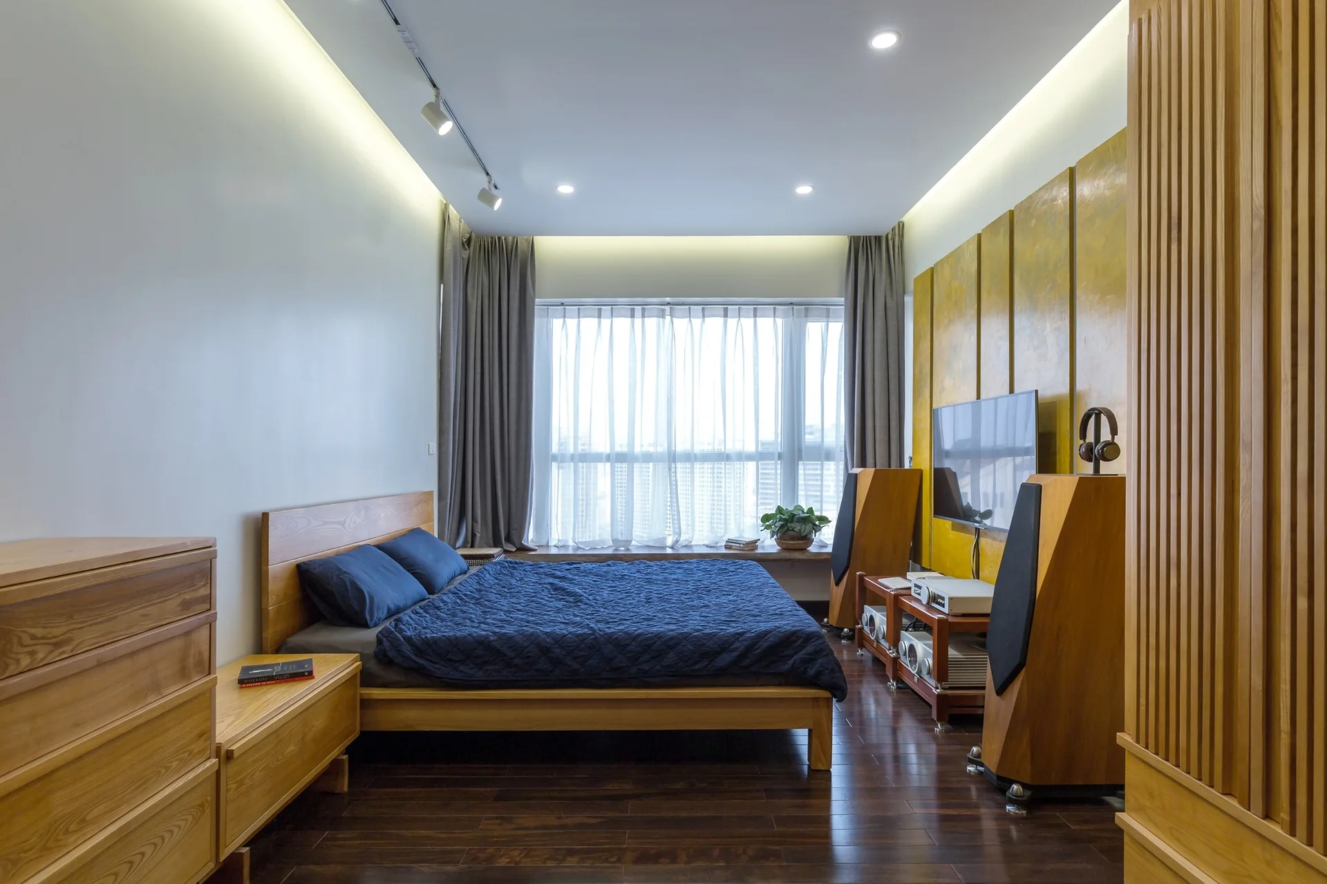 A wooden bed with deep blue sheets faces a television mounted on a textured yellow wall panel next to large wooden speakers.
