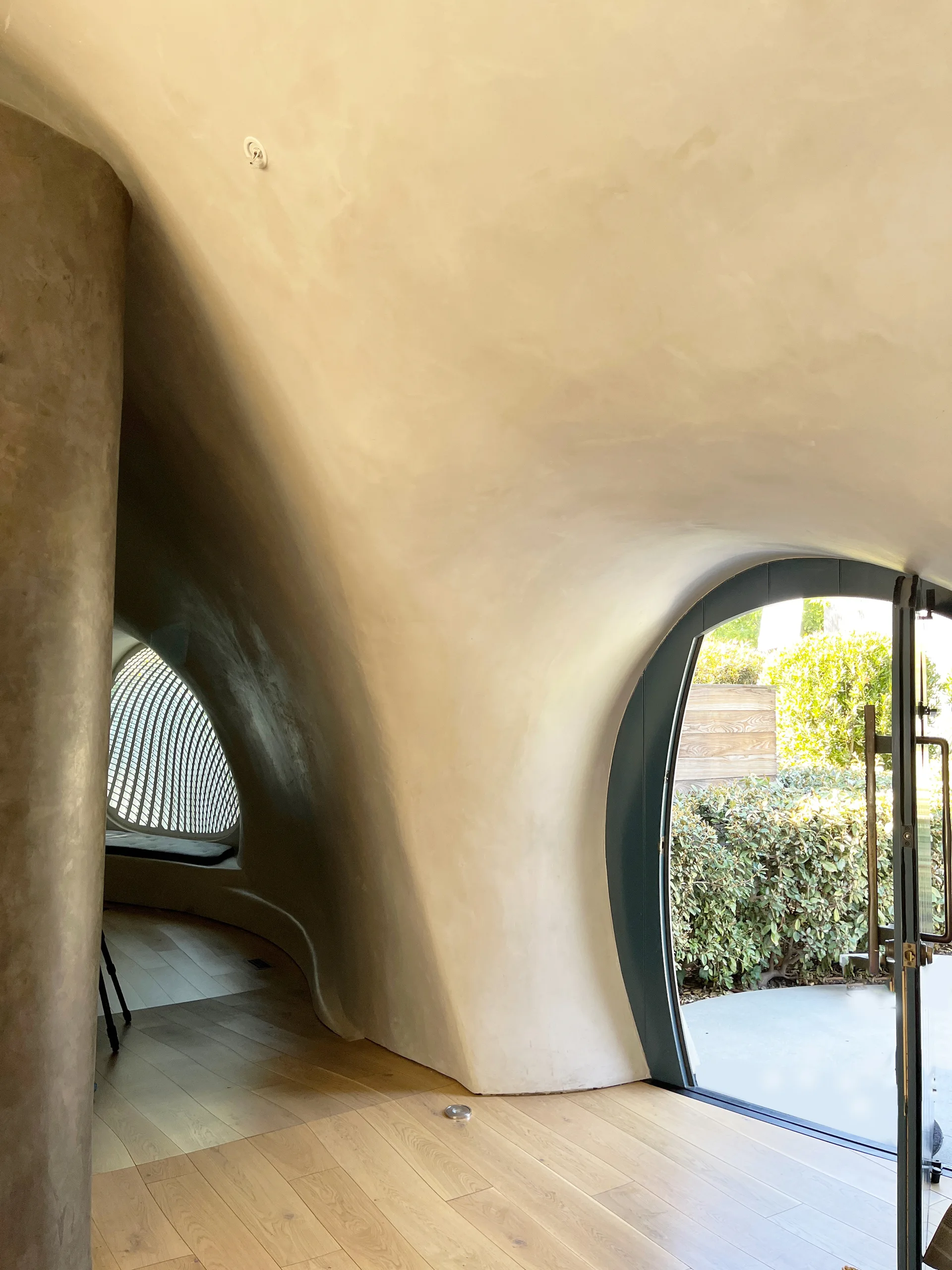 An organic interior hallway featuring sculptural curved cream plaster walls, light wood flooring, and an arched glass door opening to an outdoor patio.