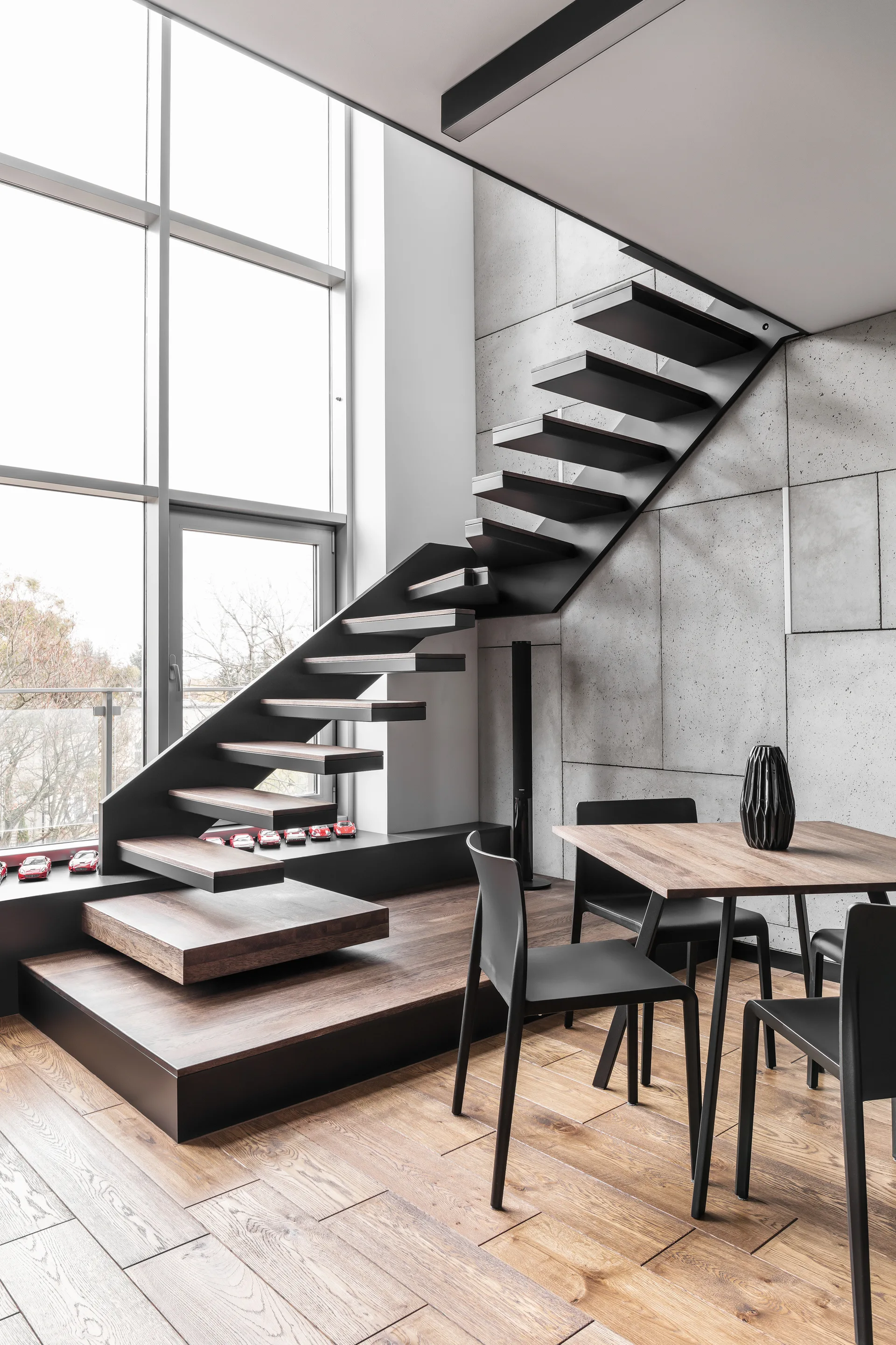 A modern dining room features a floating steel staircase with wood treads against a large concrete panel wall next to a wood table and black chairs.