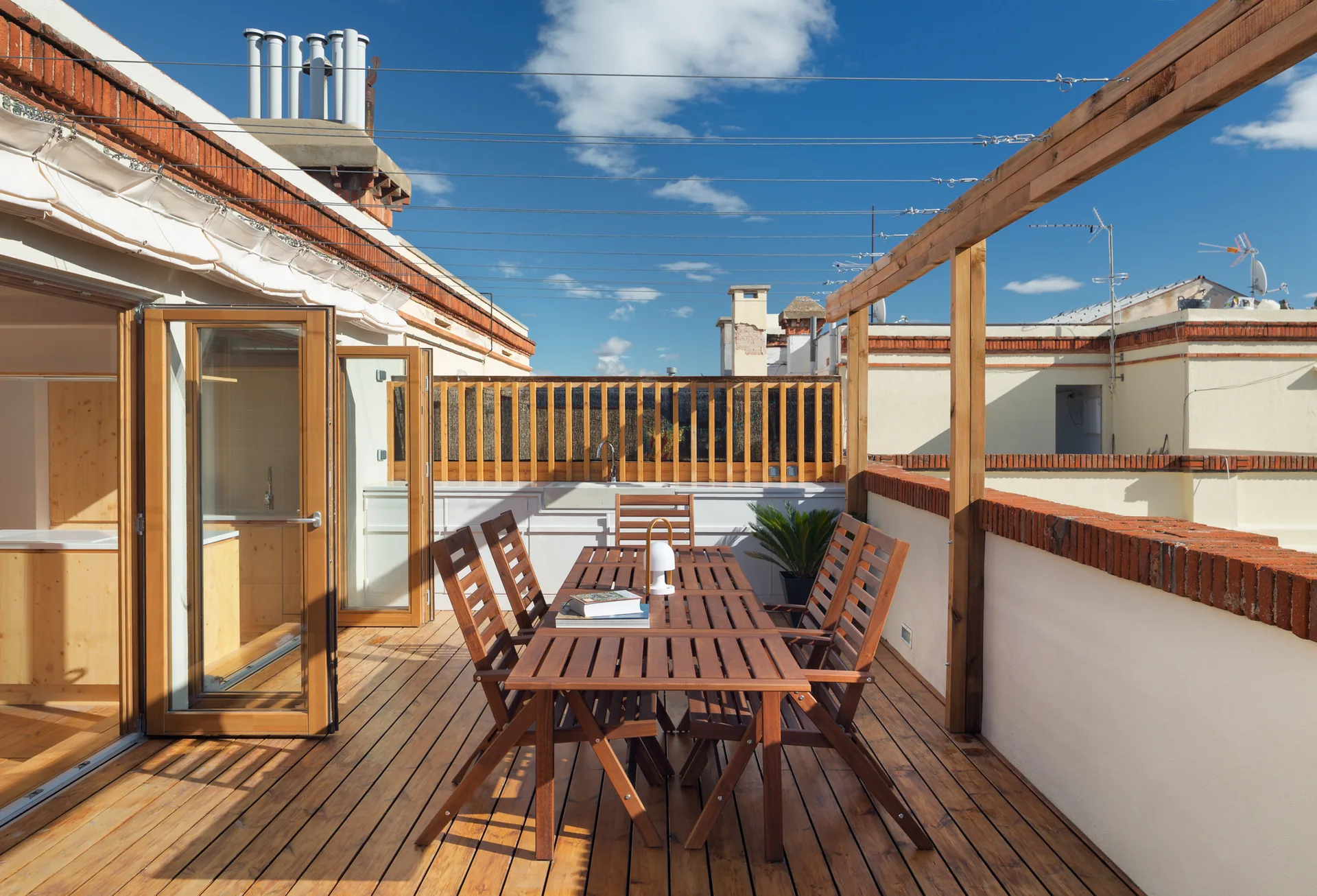 A sunny rooftop terrace features a wooden dining table and chairs on a wood deck, bordered by white walls with brick trim under a blue sky.