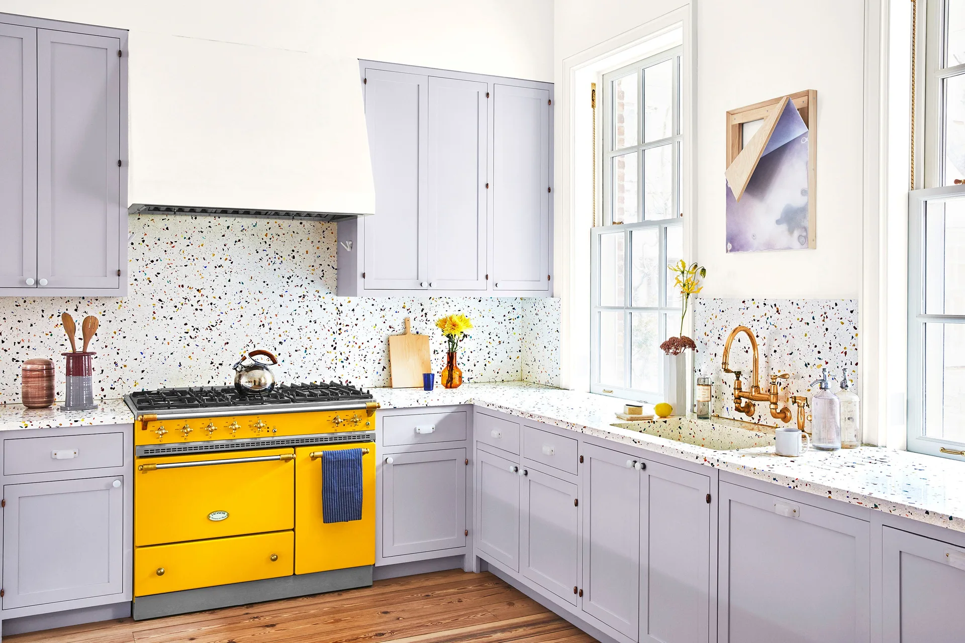 A bright kitchen features light lavender cabinets, terrazzo countertops and backsplash, and a bold yellow stove.