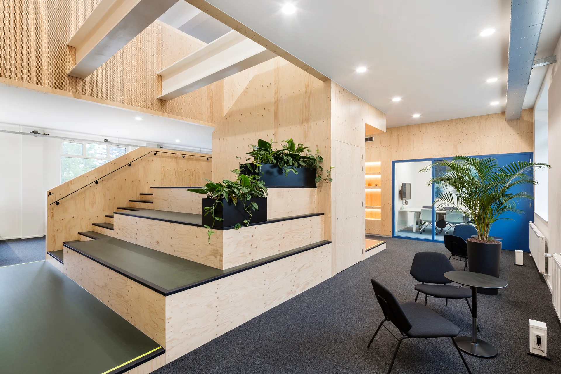 A modern tiered plywood staircase with built-in black planters and green plants sits near a grey carpeted lounge area with dark chairs.
