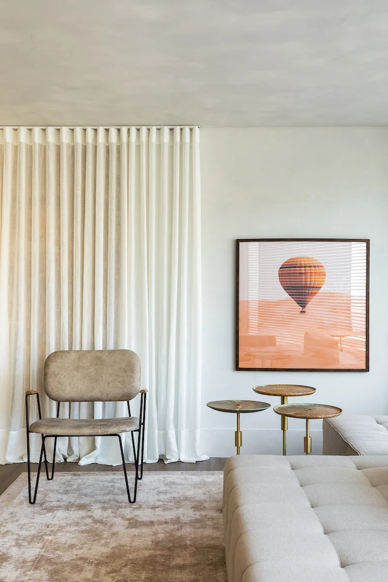 An armchair and tufted sofa sit in a minimalist living room with floor-to-ceiling white drapes and three brass nesting tables.