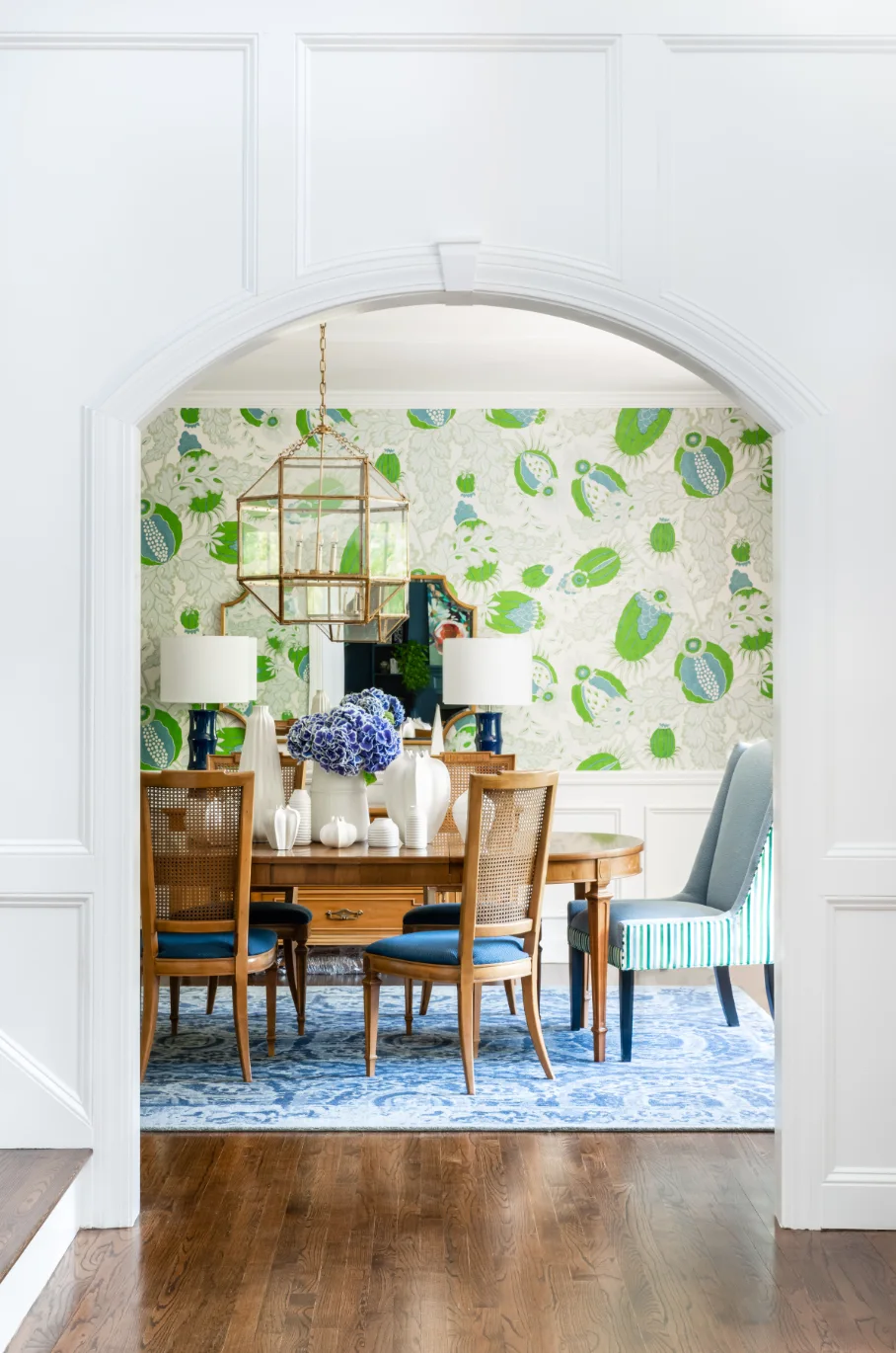 A dining room viewed through a white archway featuring bold green botanical wallpaper, a wooden table with cane-back chairs, and a blue rug.