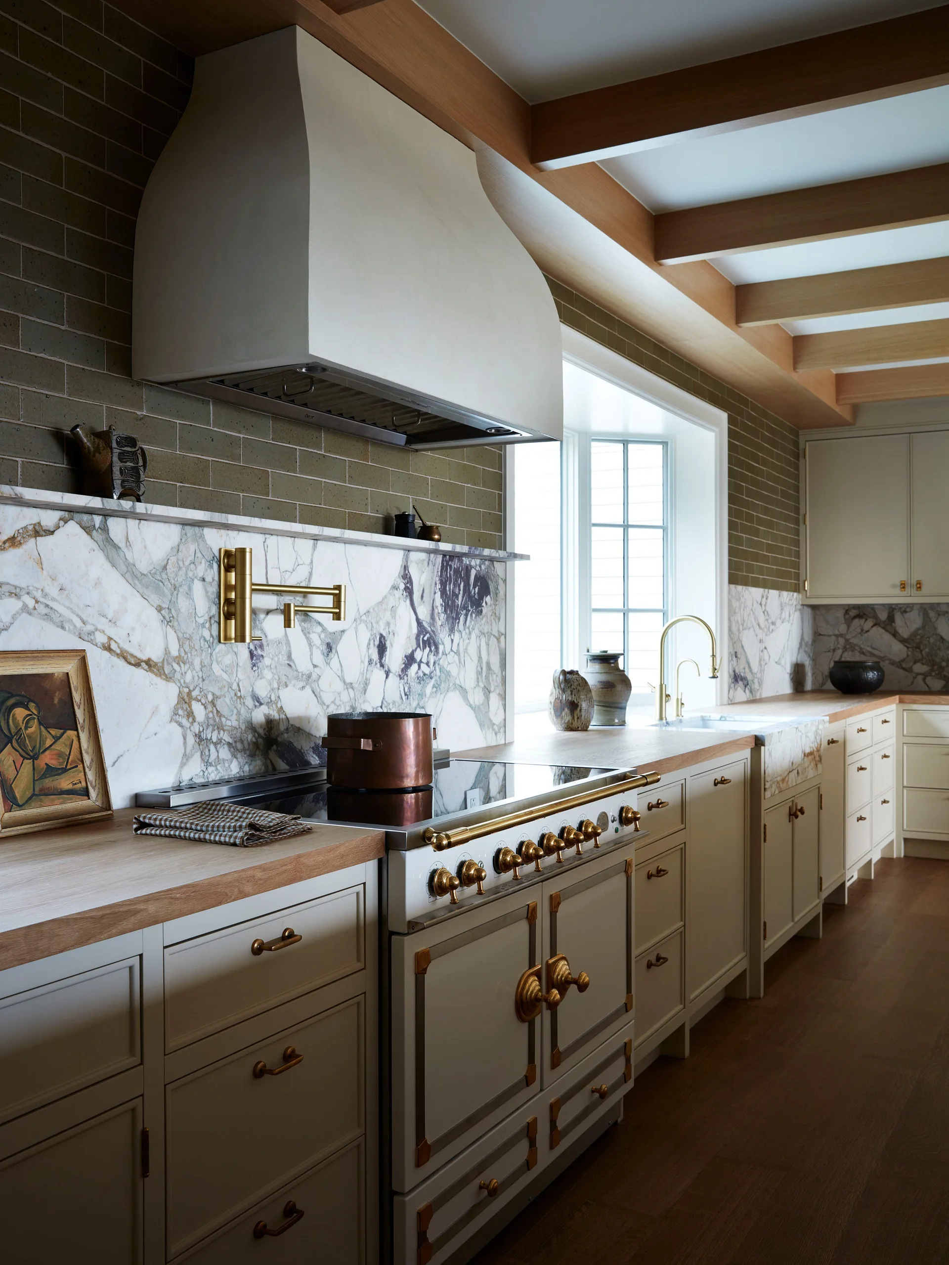 A traditional kitchen featuring cream cabinets, a custom white range hood, a marble slab backsplash, and exposed wooden ceiling beams.
