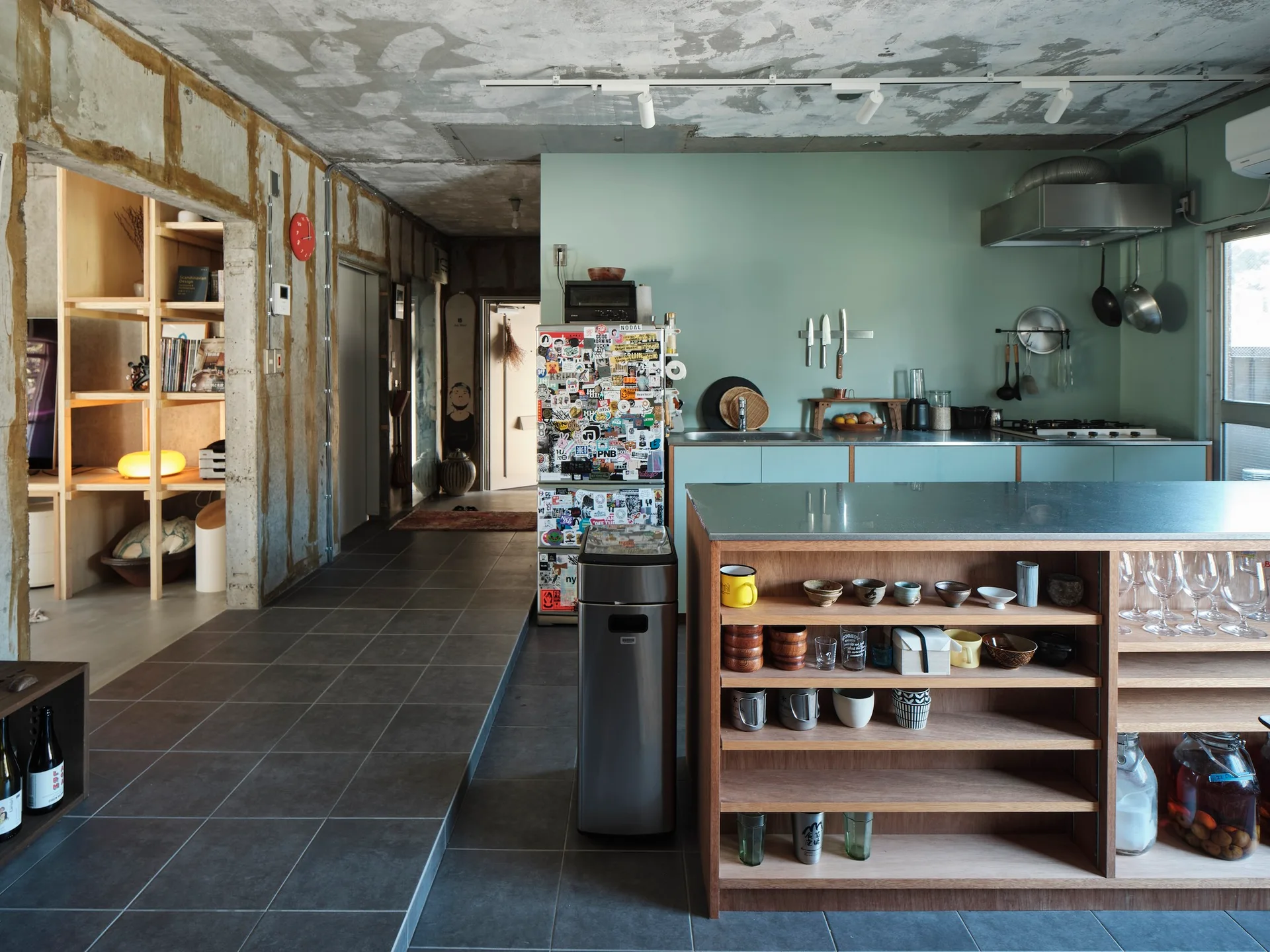 An industrial kitchen features a wooden island with open shelving, a sticker-covered refrigerator, a mint green back wall, and an exposed concrete ceiling.