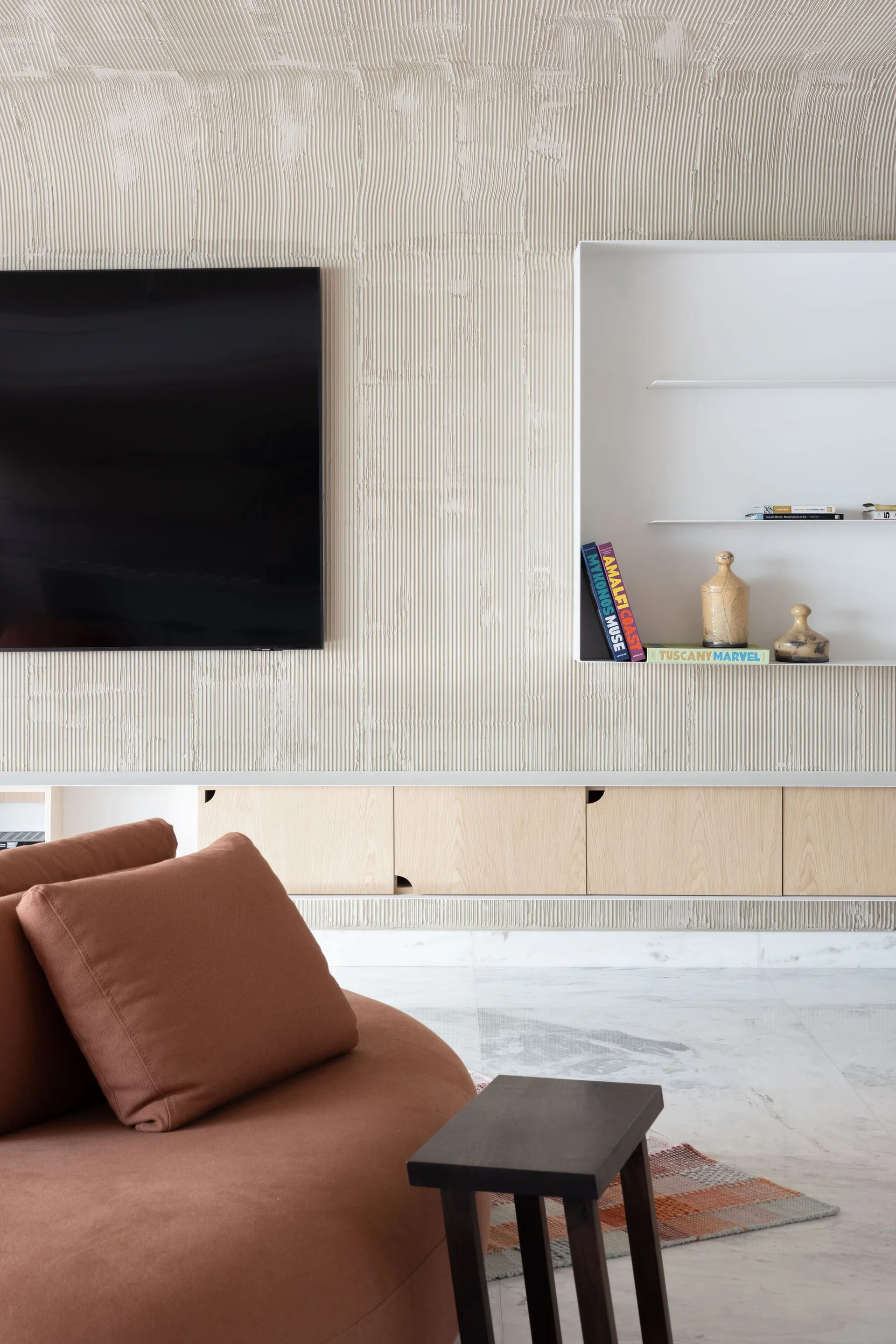 A modern living room features a vertically ribbed beige plaster wall with a mounted TV, built-in white shelving, and a curved terracotta sofa.