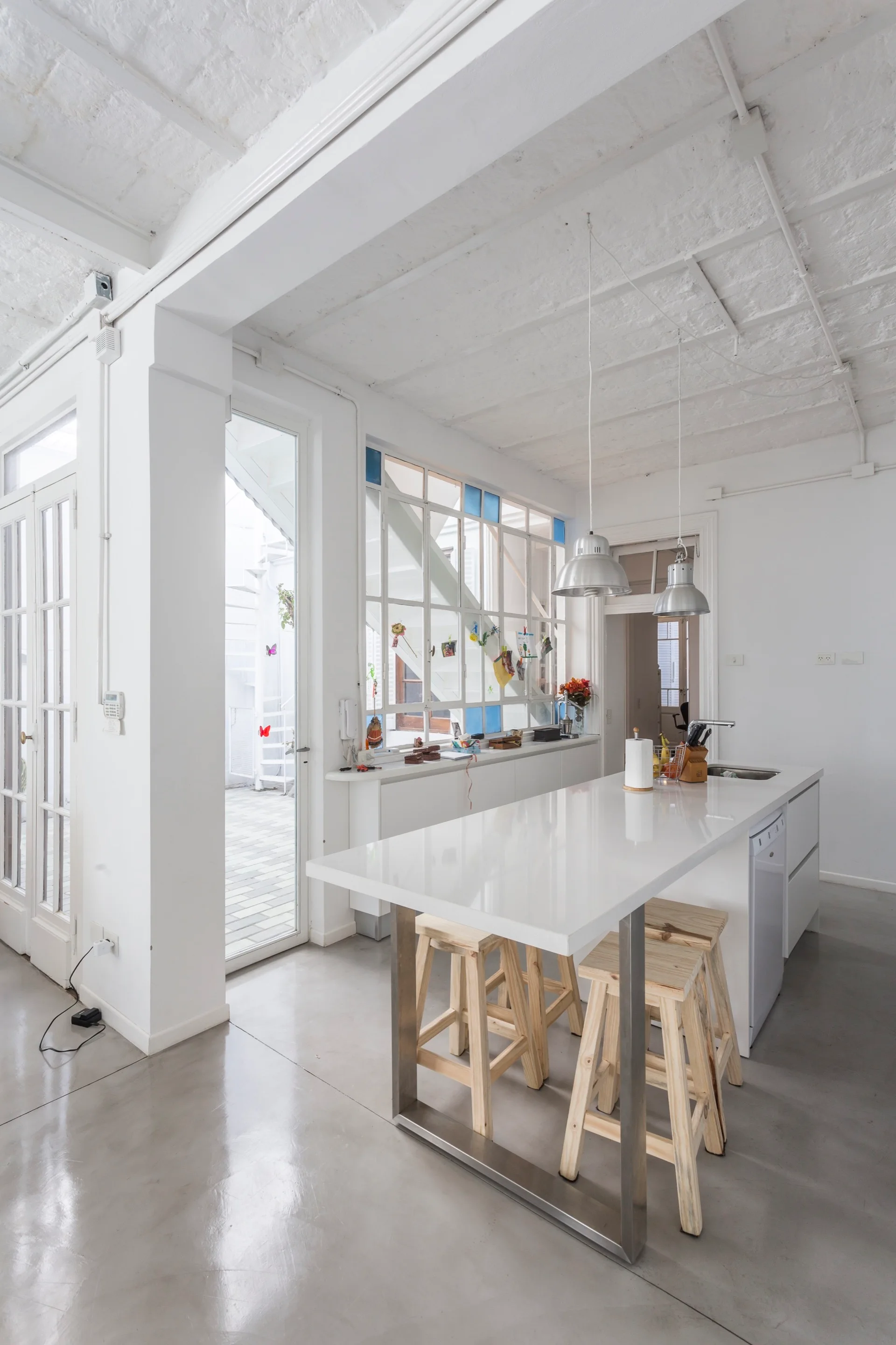 A bright kitchen features a white island with light wood bar stools, polished concrete floors, and metallic pendant lights below an exposed ceiling.