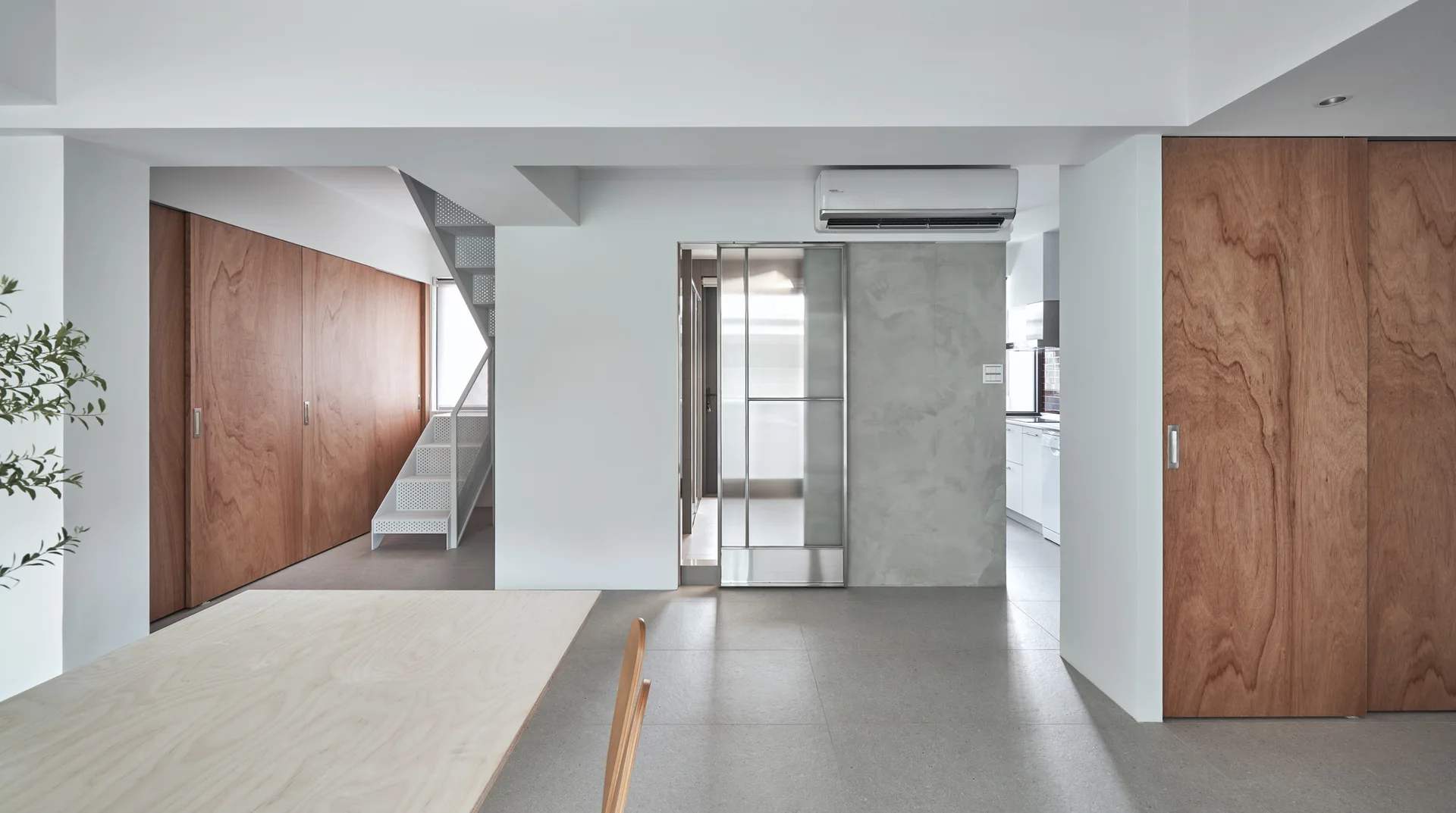 A minimalist residential interior featuring a light wood dining table, large wood-panel sliding doors, grey tile flooring, and a white staircase.