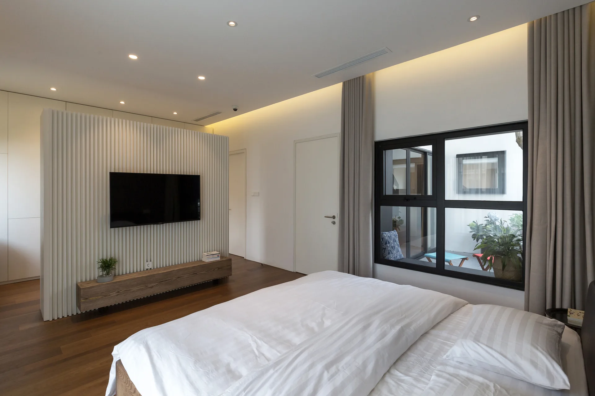 A minimalist bedroom features a bed with white bedding, a TV mounted on a white slatted partition wall, and a window viewing a small courtyard.