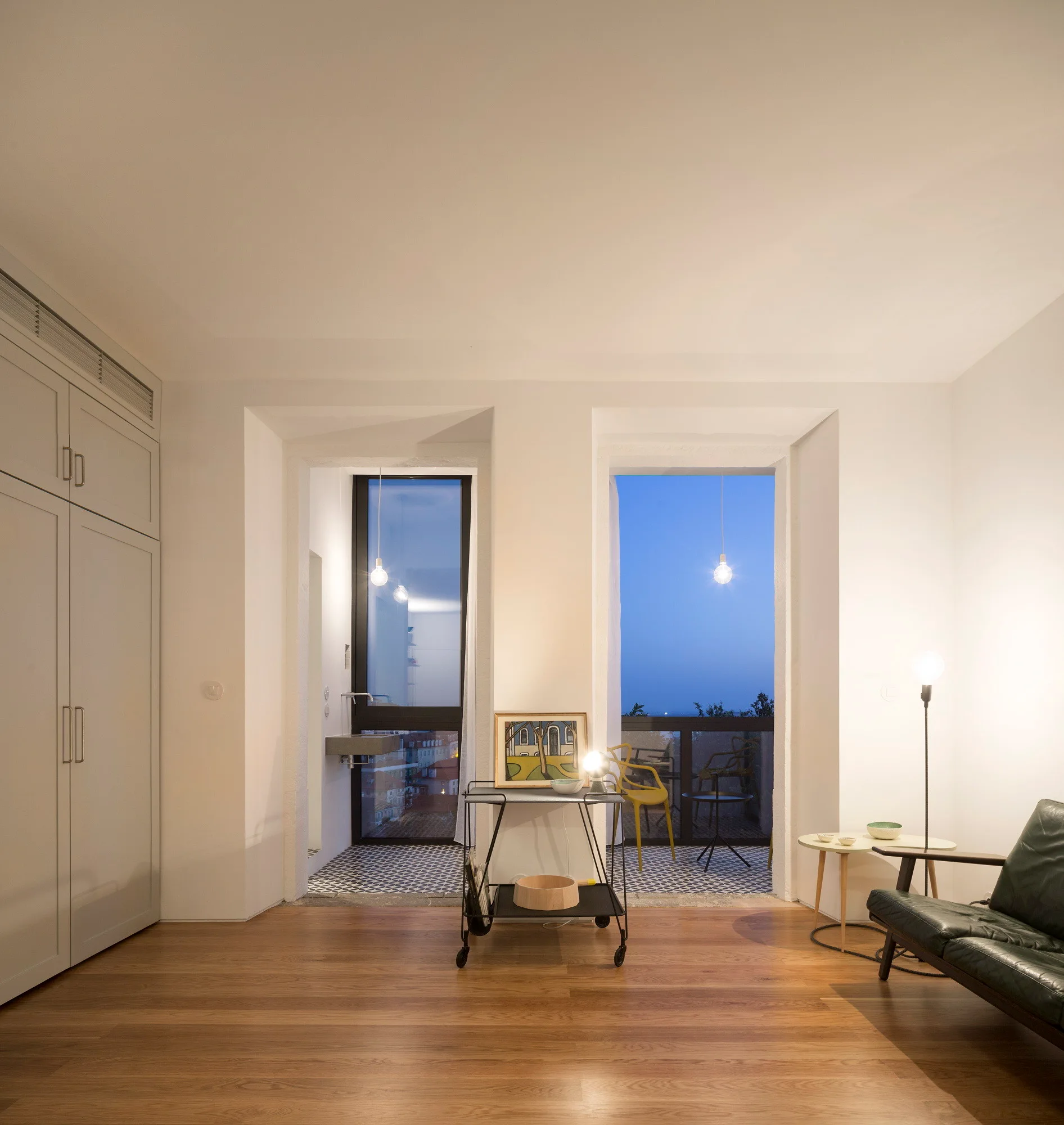 A minimalist living room featuring warm wood floors, white built-in cabinetry, and twin doorways opening onto a tiled balcony at twilight.