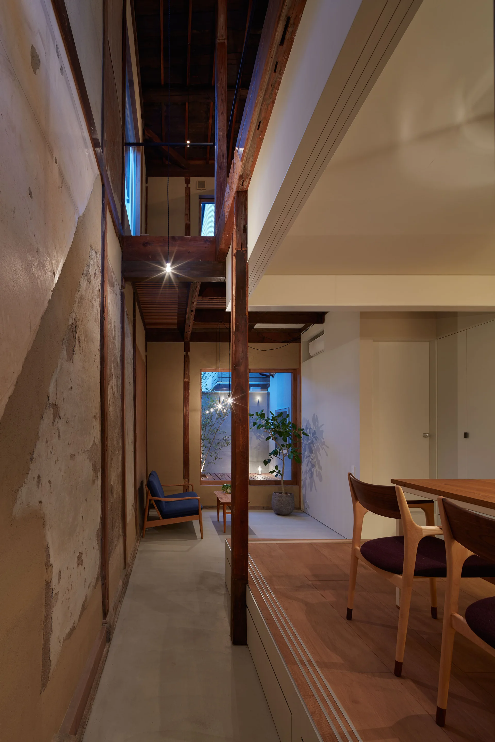 A Japanese modern split-level interior features a concrete walkway with an armchair, a raised wood dining platform, and exposed timber beams.