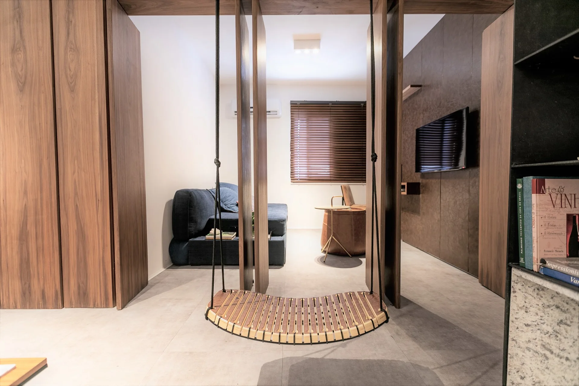 A contemporary living room featuring a curved wooden hanging swing in the foreground, wood panel dividers, and a blue sofa in the background.
