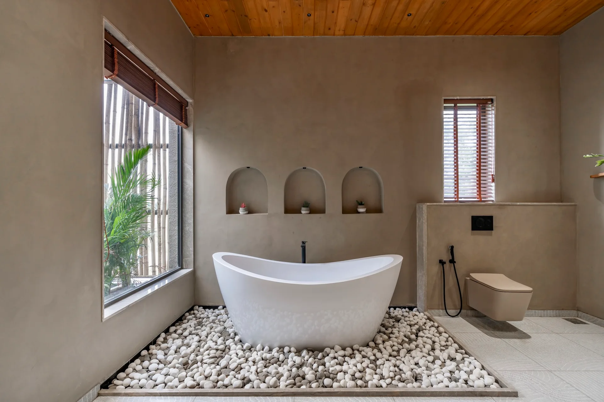A modern bathroom with a freestanding white tub resting on a bed of white pebbles, beneath smooth plaster walls with three arched niches.
