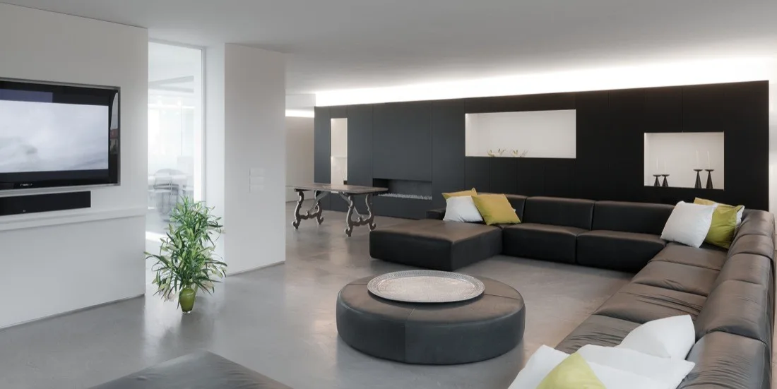A minimalist living room features a large black leather sectional sofa, a round ottoman, and a matte black wall with integrated lighting and niches.