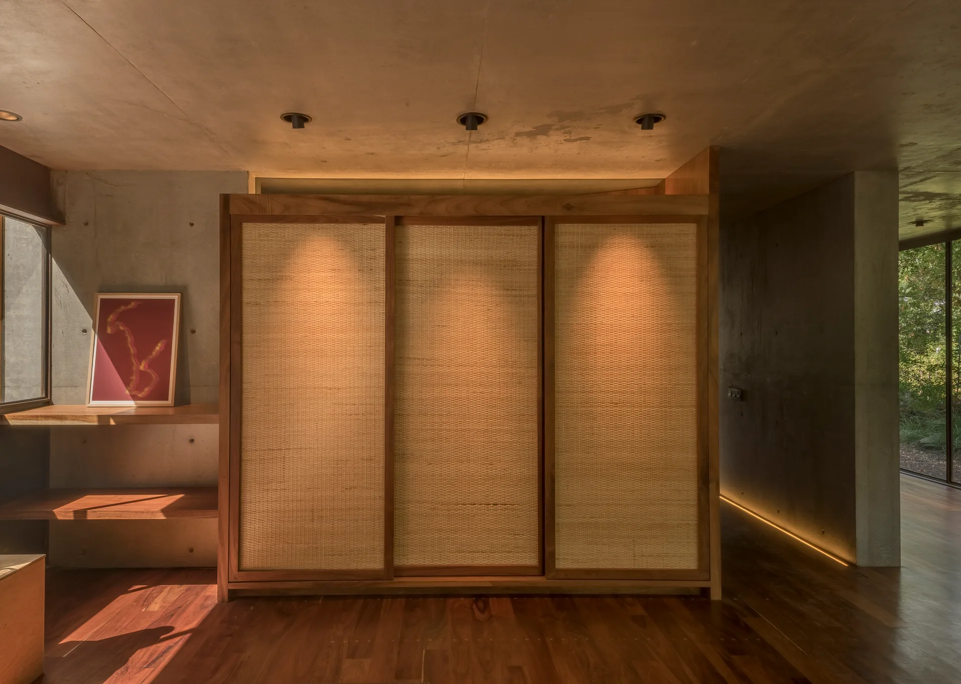 A large wooden wardrobe with woven cane sliding doors stands in a minimalist room with exposed concrete walls and hardwood floors.